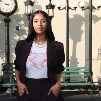 Woman in a blazer stands by a green bench and ornate street lamps near a vintage clock, evoking Main Street USA at Disneyland.
