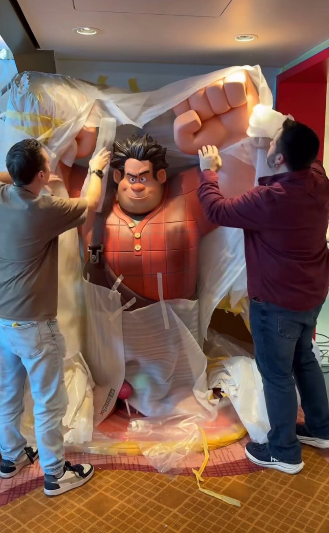 Two people unveil a Wreck-It Ralph statue bursting from packaging, echoing the playful spirit seen at Disney parks worldwide.