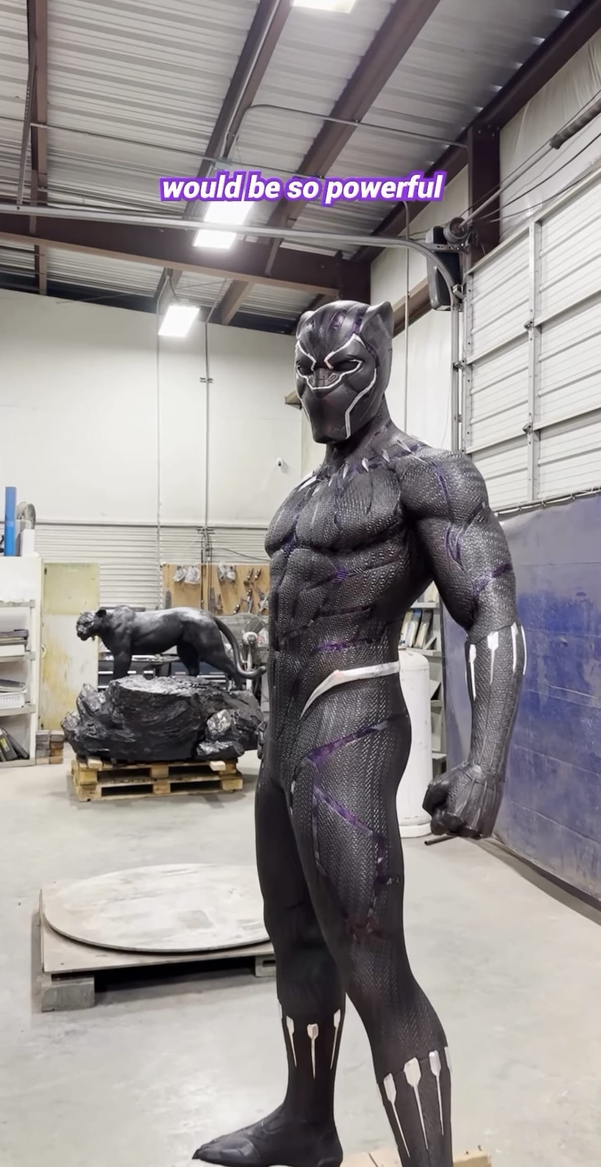 A realistic Black Panther suit display stands in a Disney workshop, with a Disney Imagineering statue in the background.