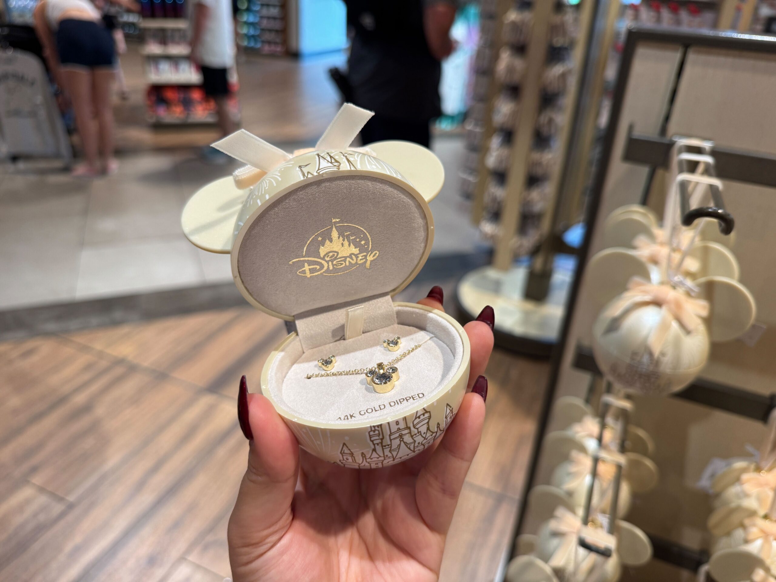Hand holding a Walt Disney World jewelry box shaped like Mickey ears, displaying a gold-dipped necklace inside a store.