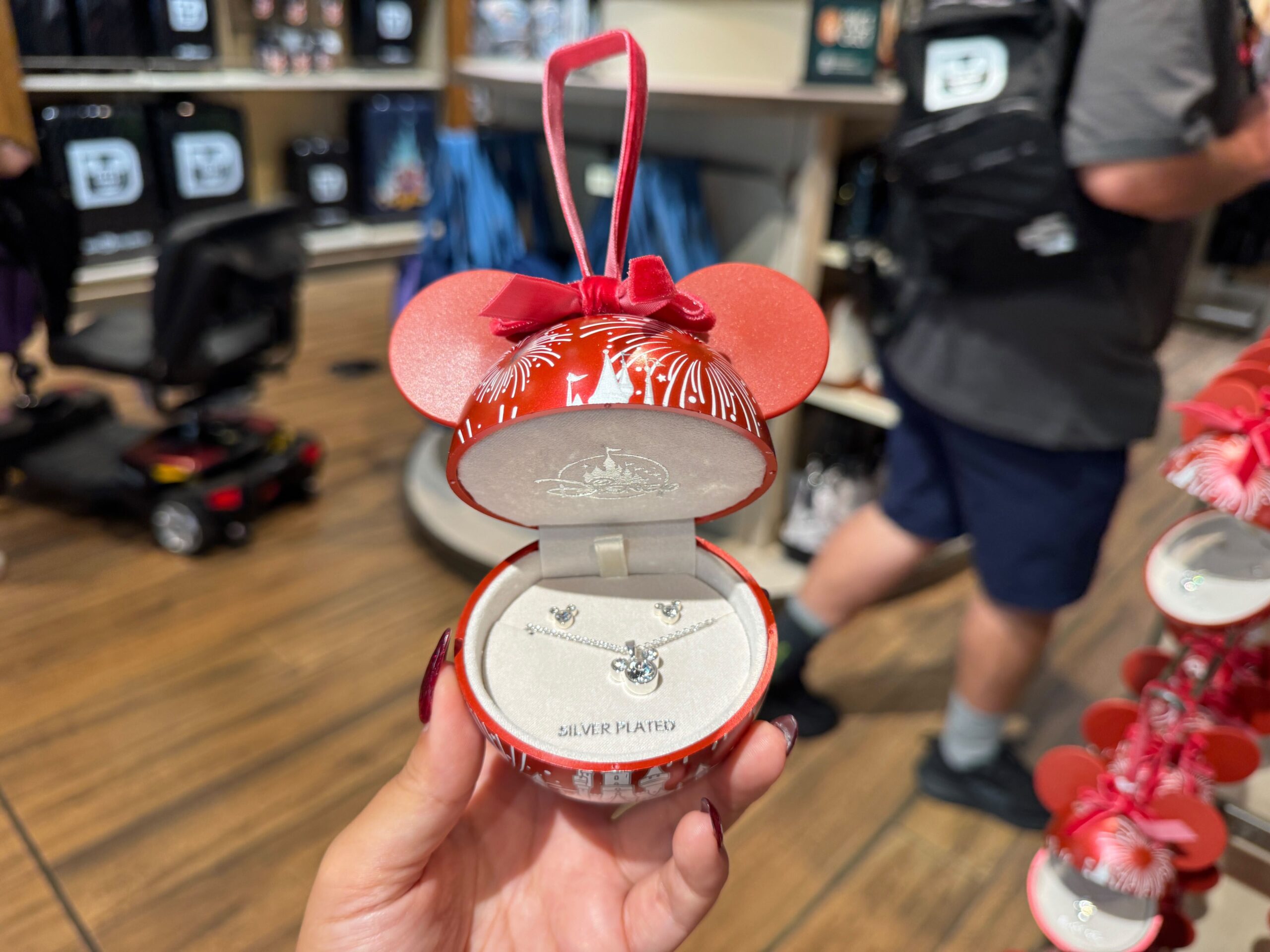 A hand holds a red Walt Disney World ornament box with a silver necklace and earrings set inside a store.