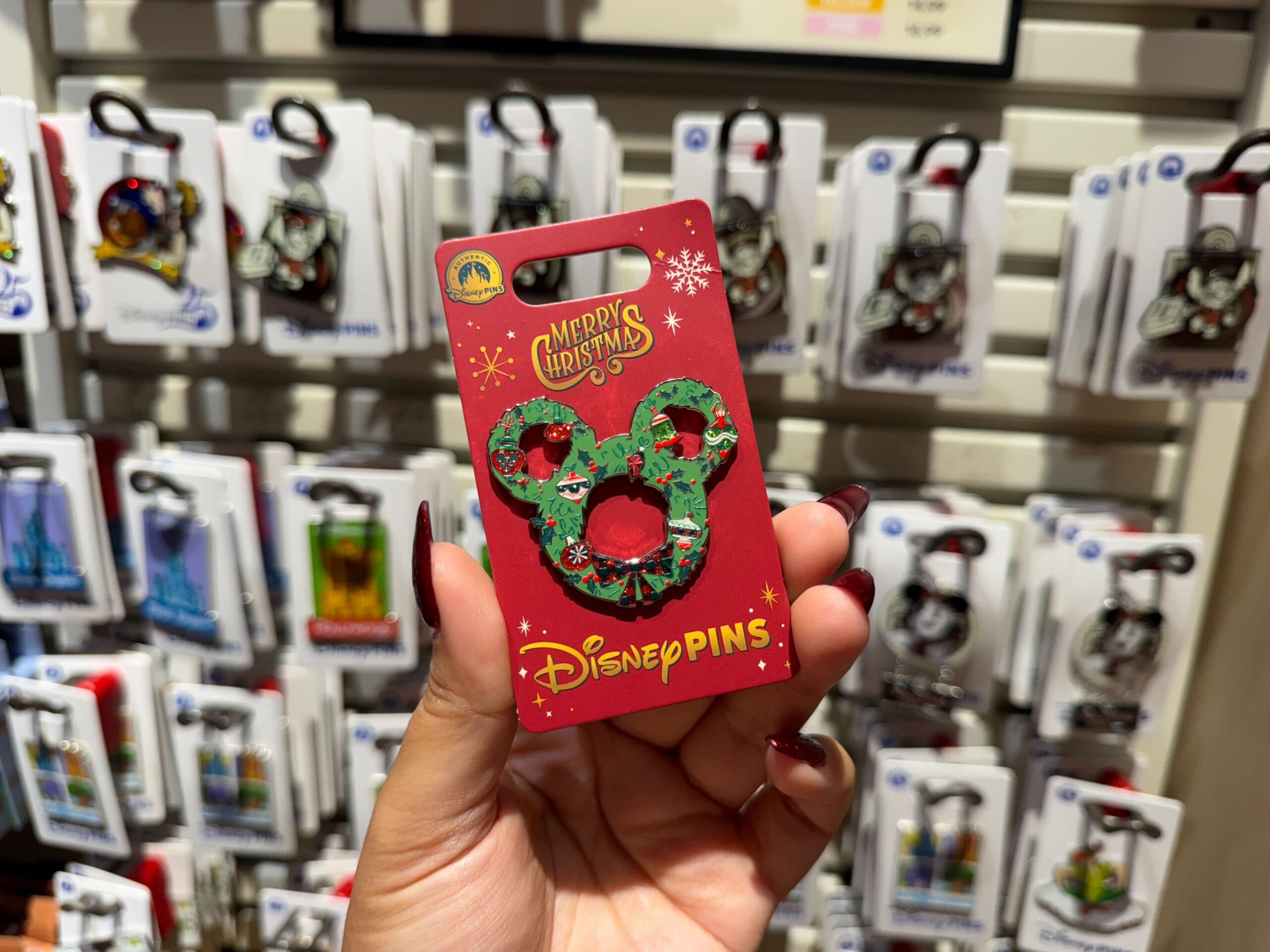 A hand holds Walt Disney World Christmas merchandise: a Mickey wreath pin, with other Disney pins displayed in the background.