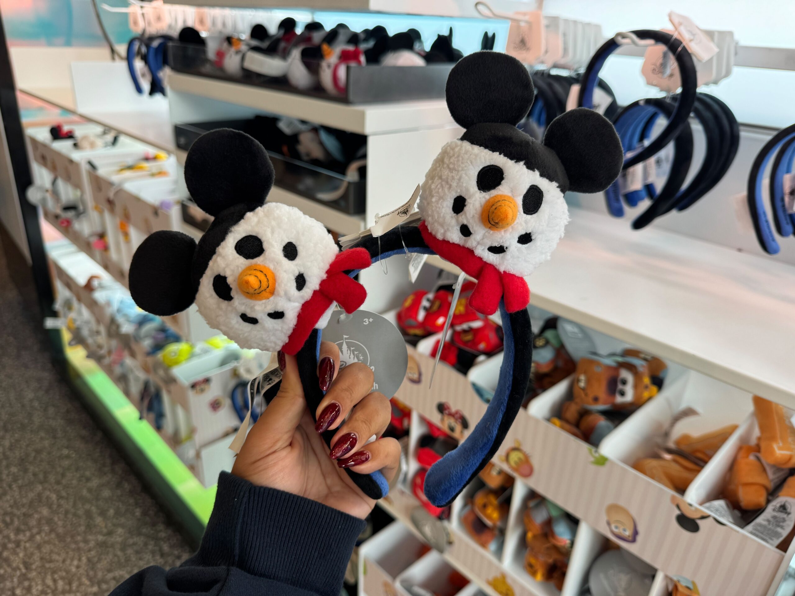 A hand holds a snowman-themed Mickey ear headband amid 2025 Disney World Christmas merchandise on display.