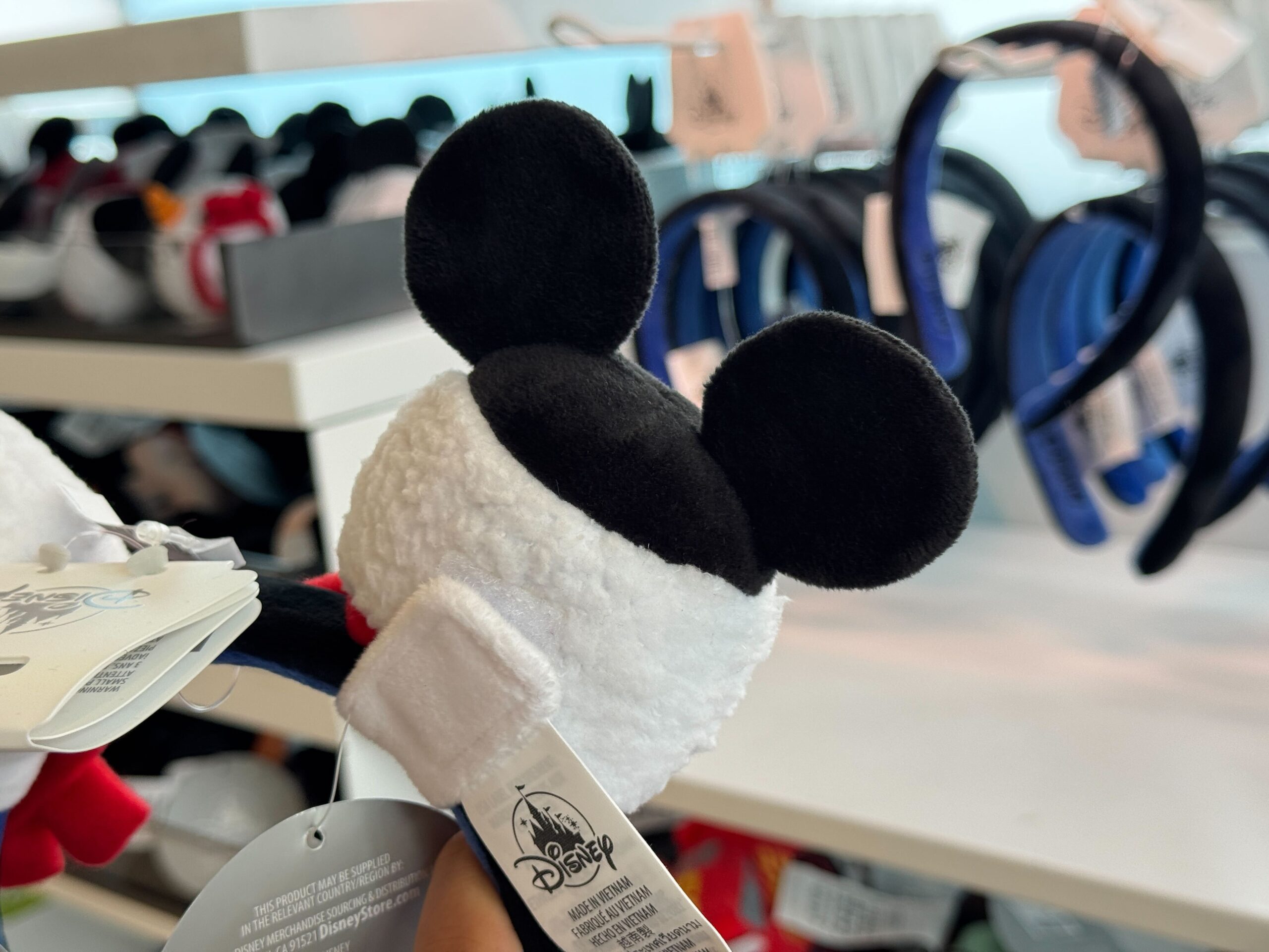 A plush Mickey Mouse keychain is held in a store aisle with 2025 Disney merchandise in the background.