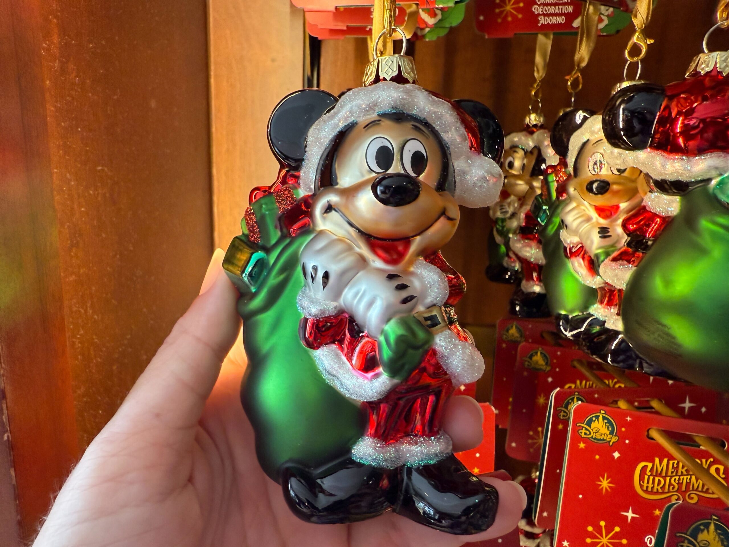 A hand holds Walt Disney World Christmas merchandise: a 2025 Mickey Mouse Santa ornament by a large green sack.
