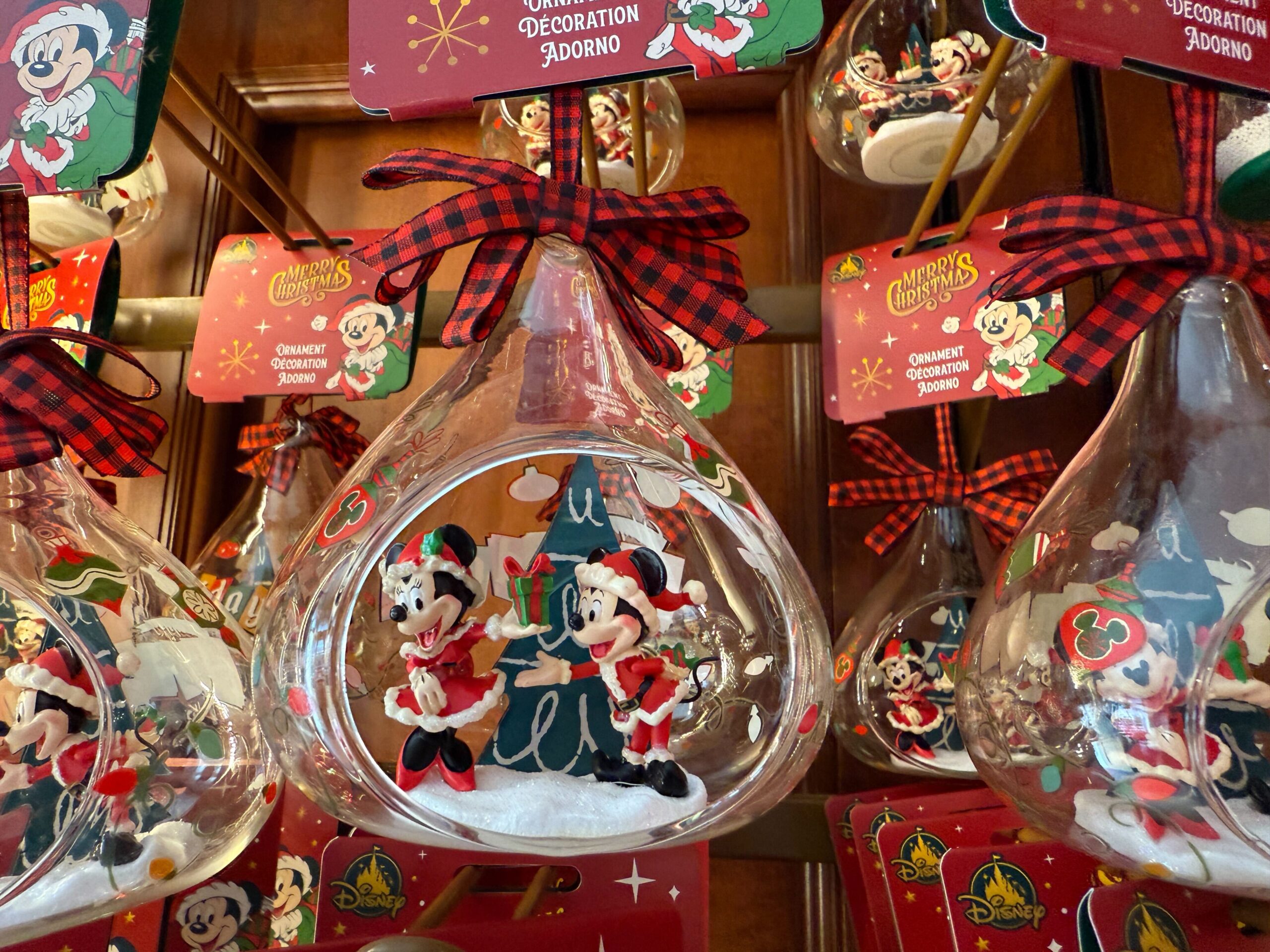 Clear glass ornament from Walt Disney World features Minnie and Mickey in festive outfits, hanging with red plaid ribbon.