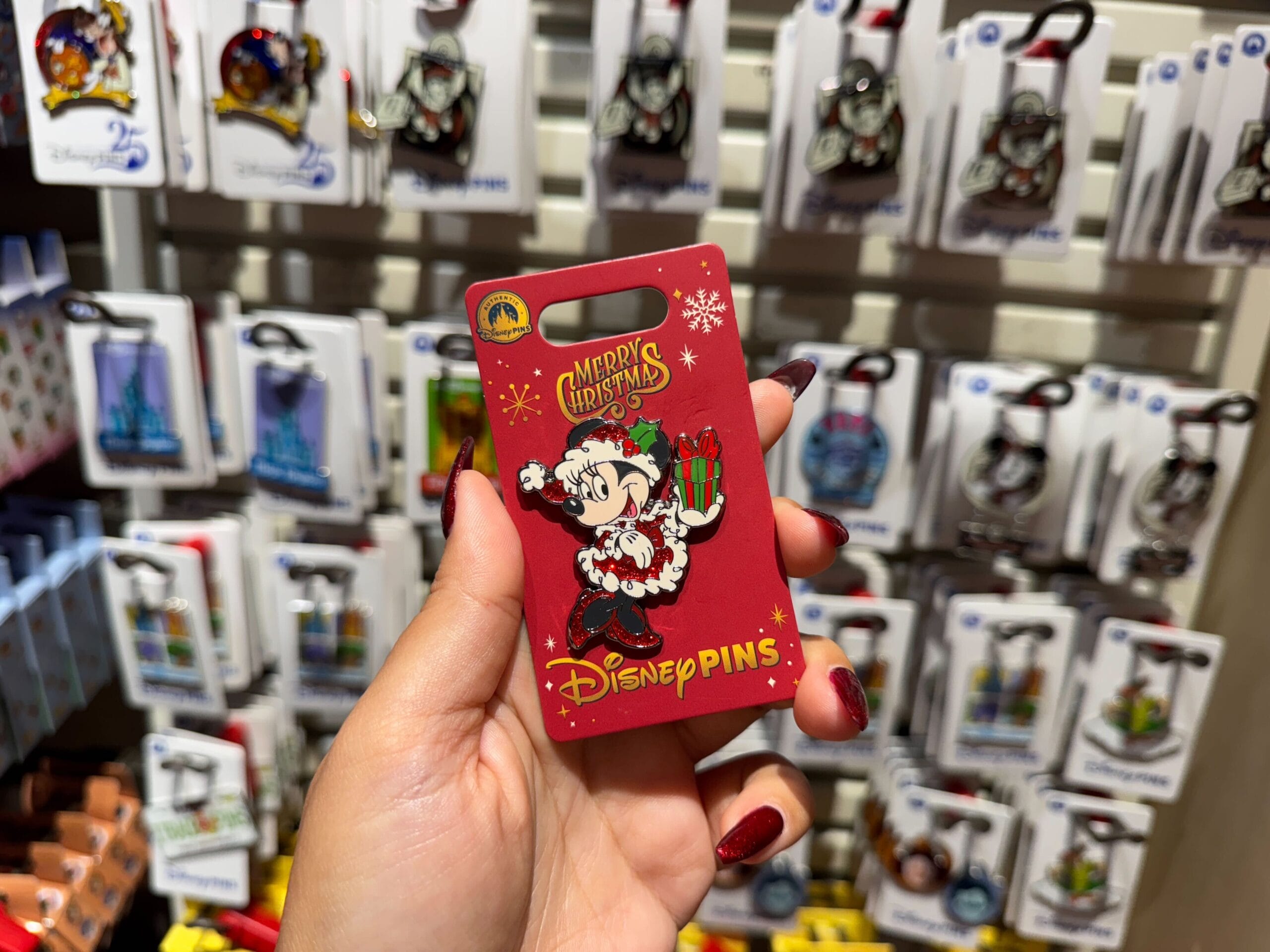 A hand holds a red Walt Disney World holiday pin featuring Minnie Mouse in front of Christmas merchandise displays.