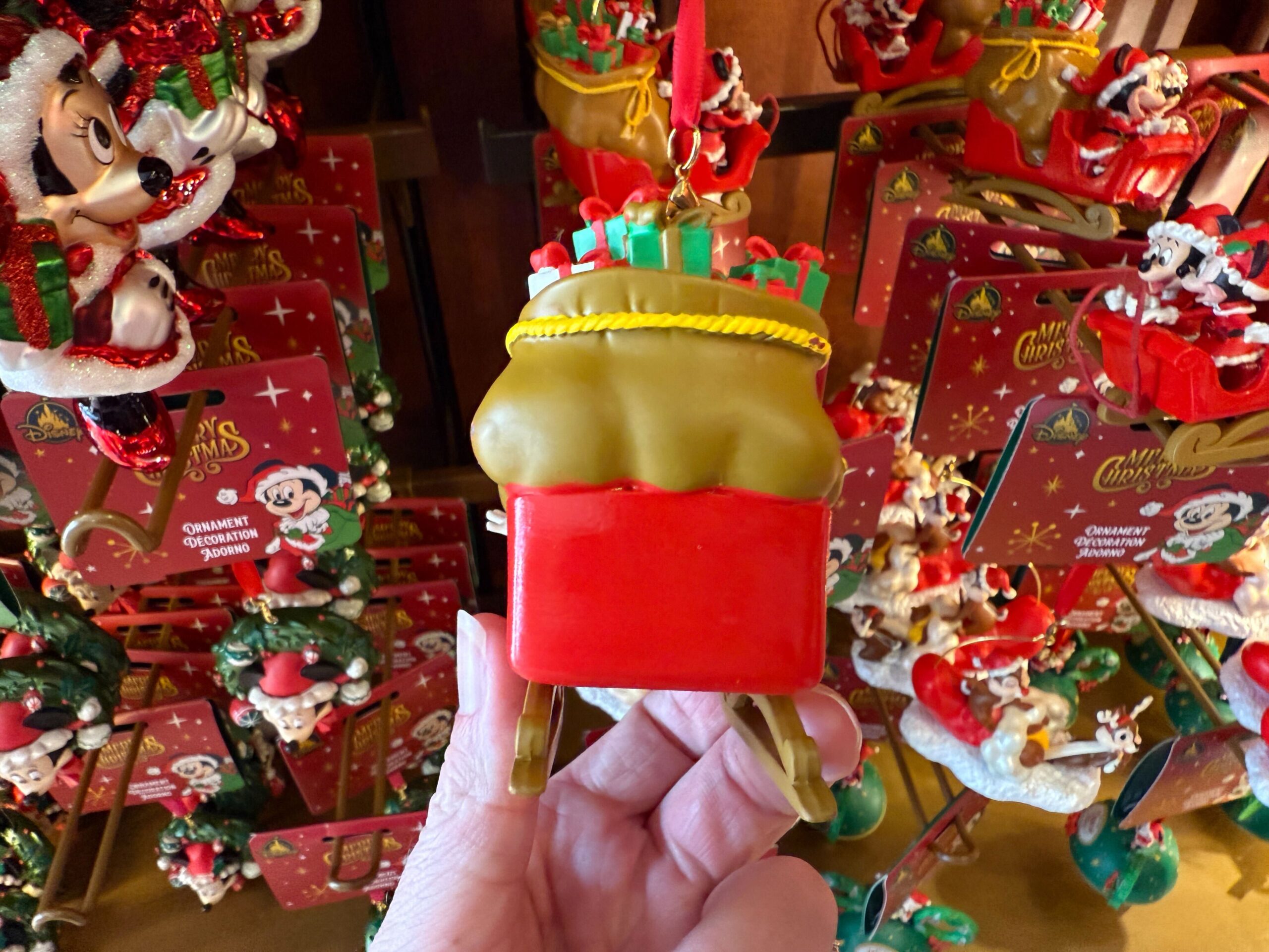 A hand holds a 2025 Christmas ornament sleigh filled with gifts, surrounded by festive Walt Disney World merchandise.