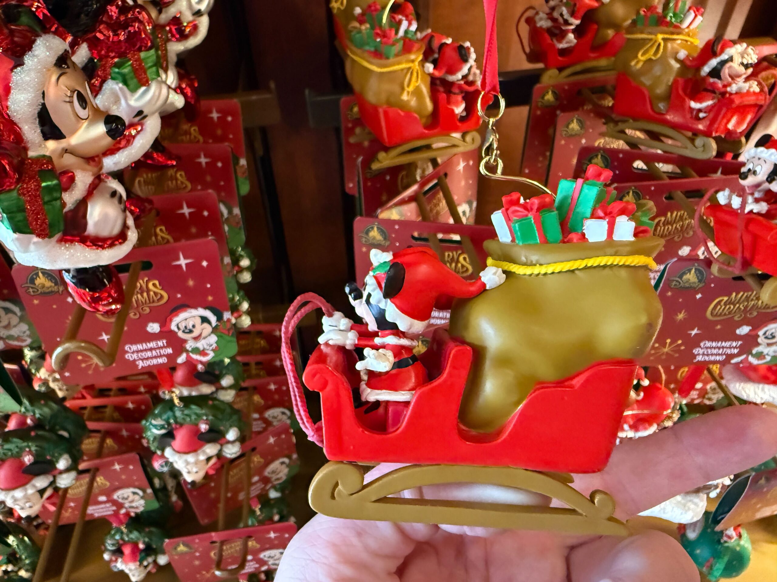 A hand holds a Walt Disney World Mickey Mouse Christmas ornament of Mickey in a red sleigh with gifts and a gold sack.