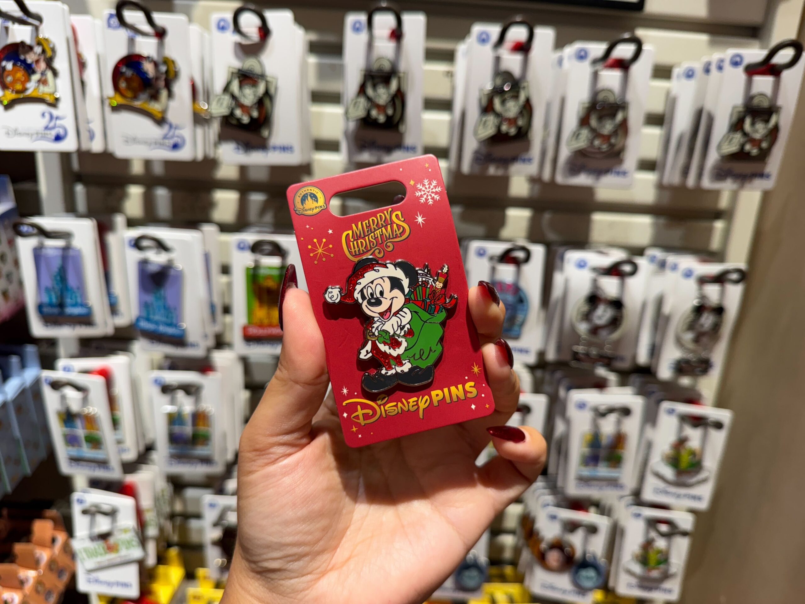 A hand holds a red Walt Disney World Christmas pin featuring Mickey Mouse in a Santa suit, with festive pins behind.