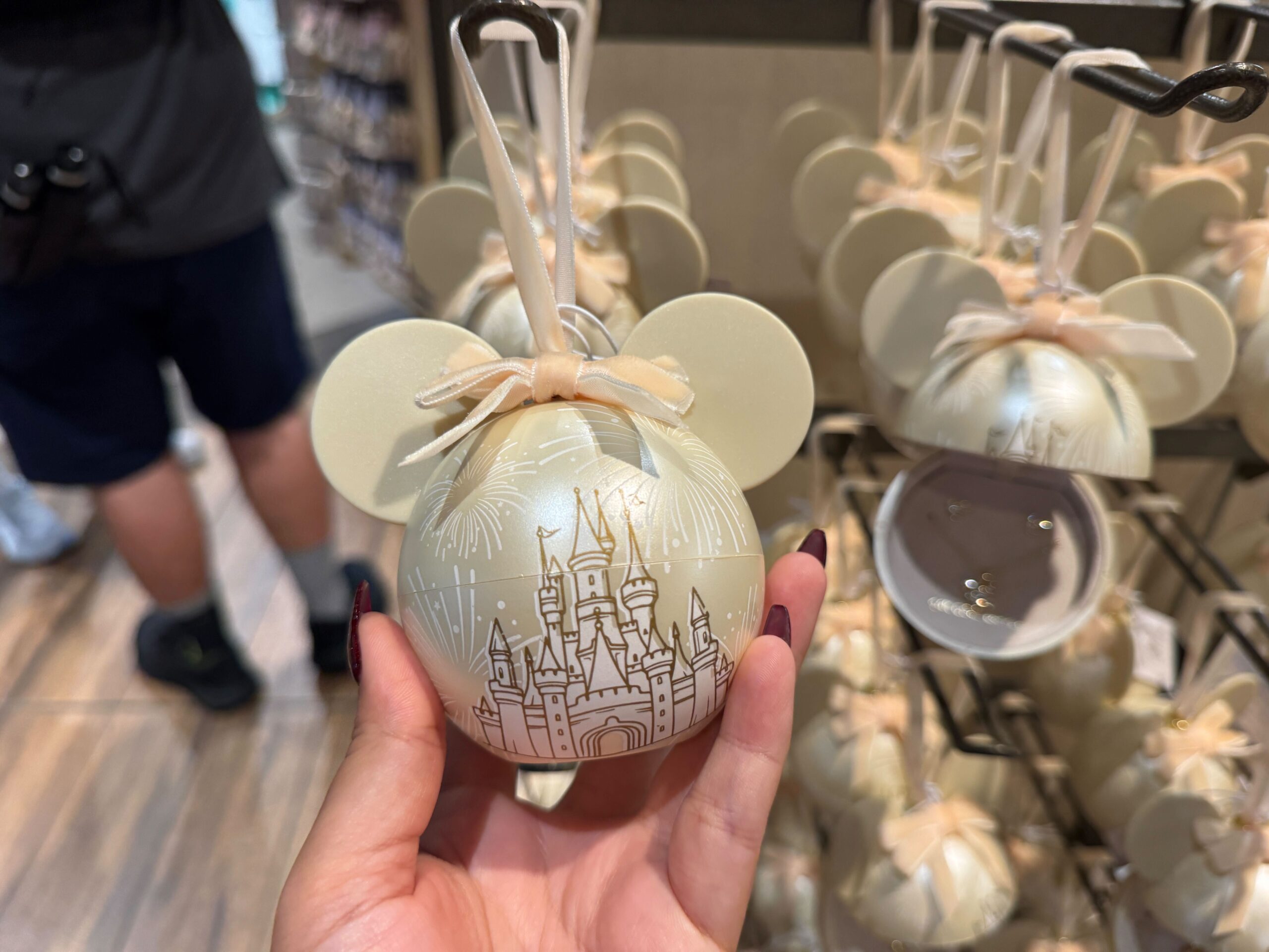 Hand holding a cream-colored Walt Disney World 2025 Mickey Mouse ornament among Christmas merchandise in a store.