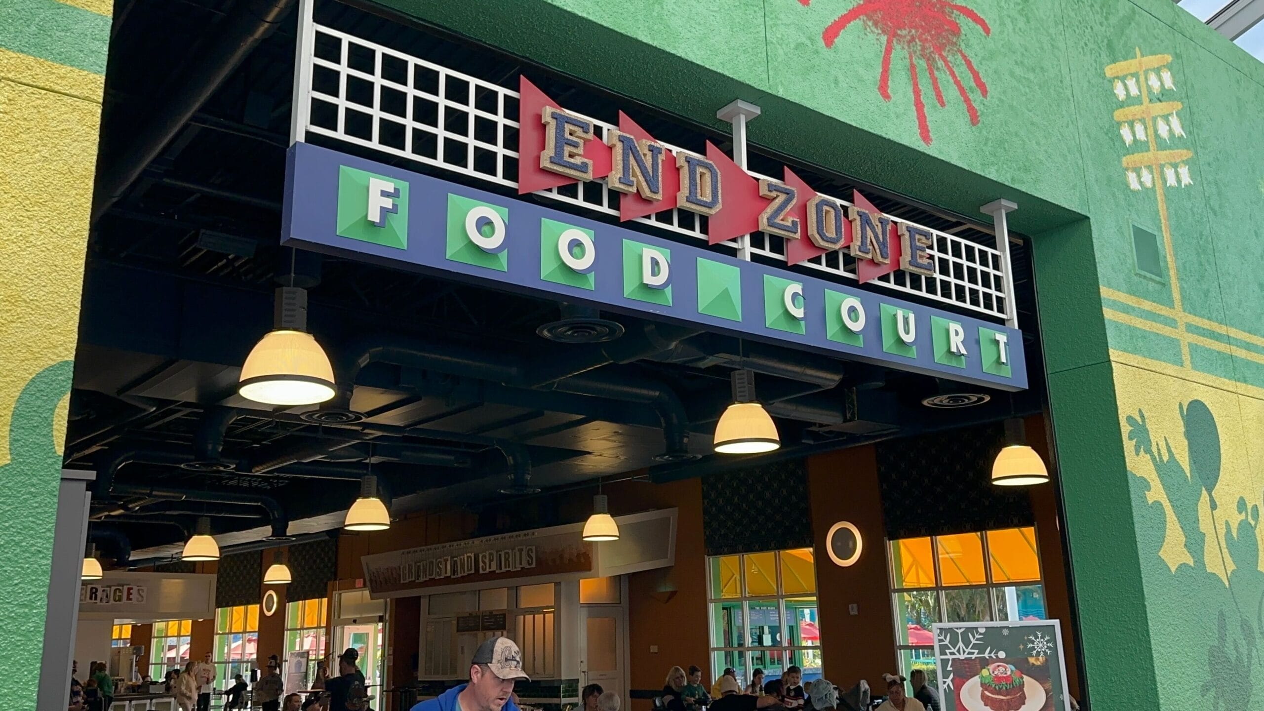 Entrance to End Zone Food Court at a Disney park, with vibrant signs, twinkling lights, and guests savoring Jerk BBQ Chicken indoors.