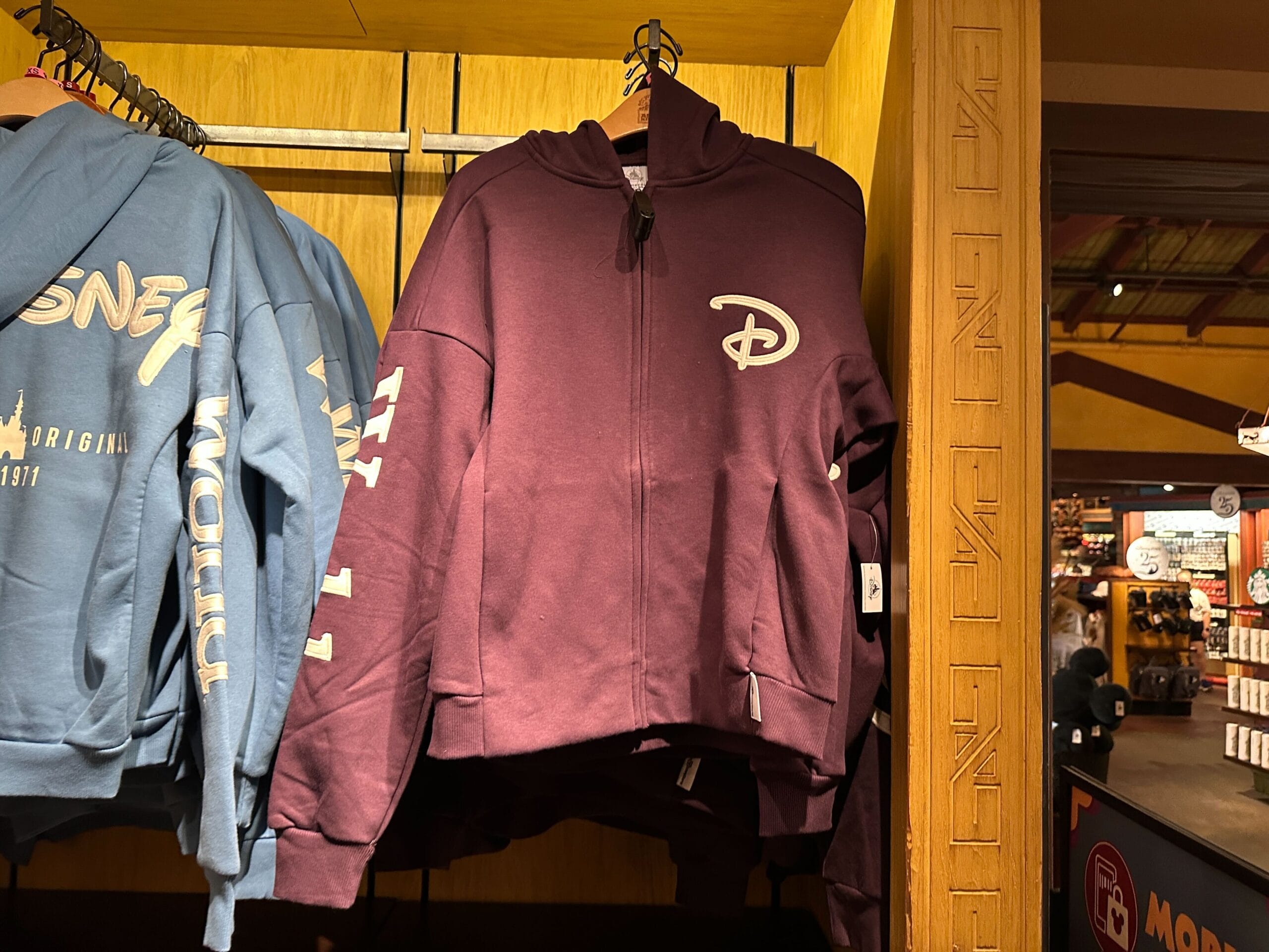 A maroon Walt Disney World zip-up hoodie hangs beside a blue Authentic Original Jacket on a merchandise rack inside the park.