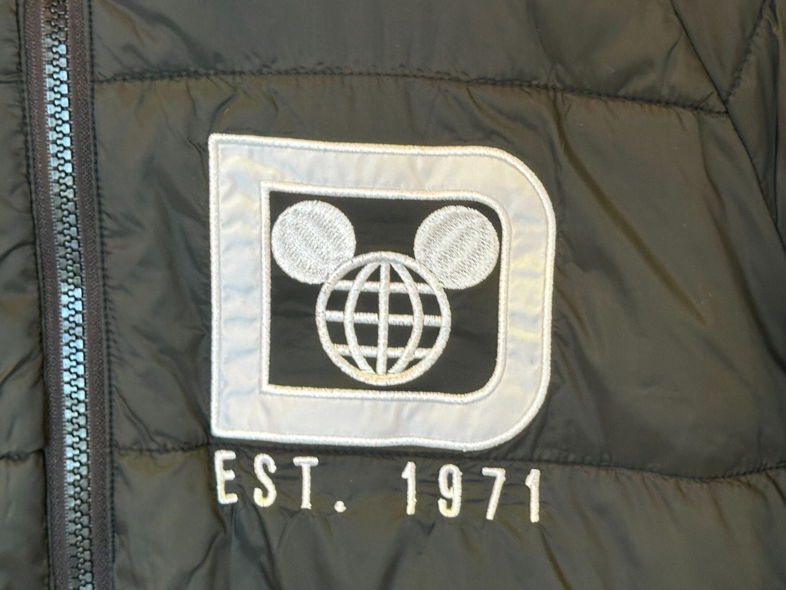 Classic black jacket with white patch featuring Mickey ears, globe, and "EST. 1971," representing Walt Disney World's legacy.
