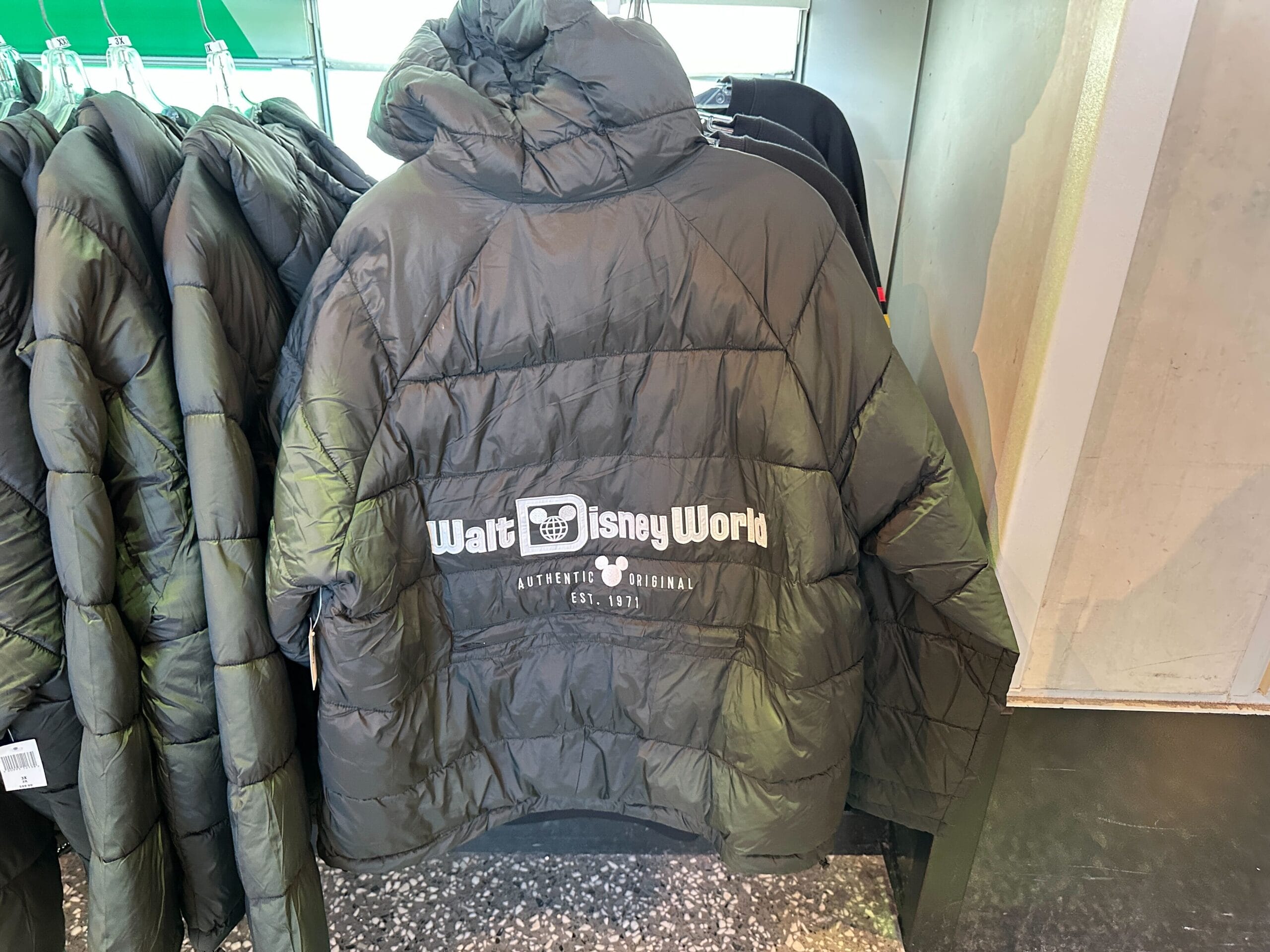 A black “Walt Disney World” Authentic Original Jacket with the classic logo on the back, displayed on a rack in a Disney Park store.