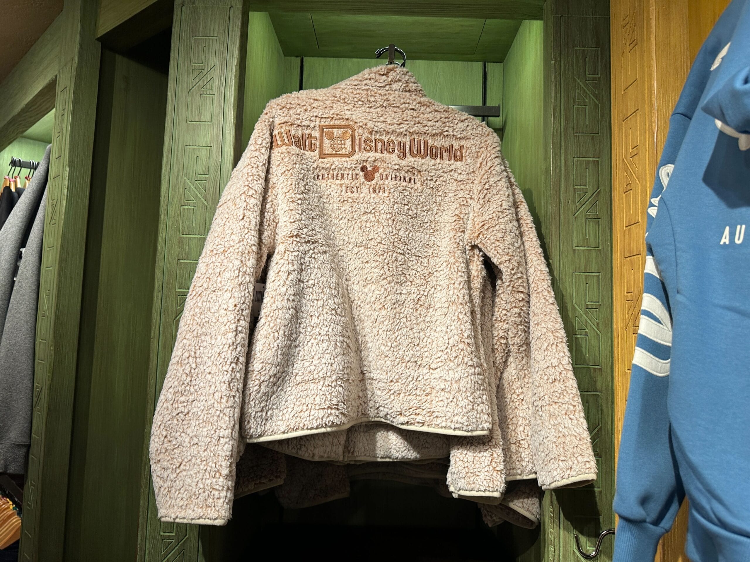 A tan sherpa jacket with "Walt Disney World" lettering hangs on a green display rack, evoking classic park merchandise style.