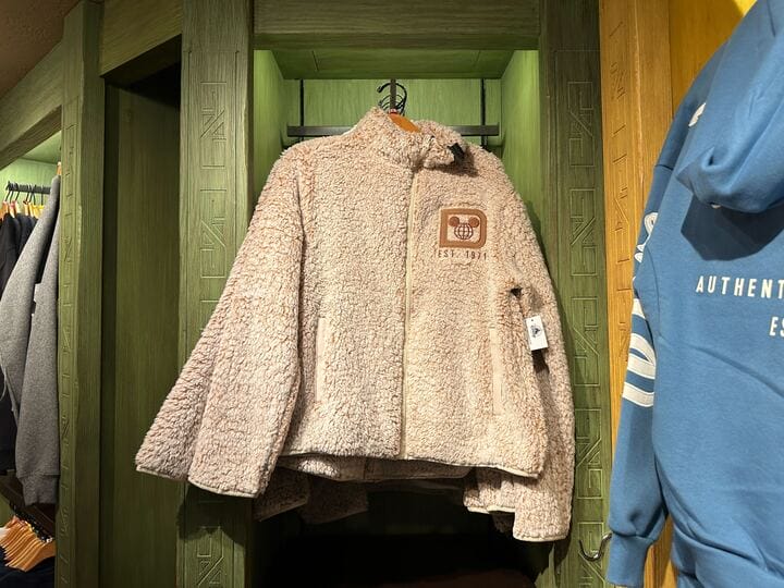 A cozy beige sherpa jacket with a Disney patch hangs on a green rack next to a blue hoodie, like merch seen at Disneyland shops.