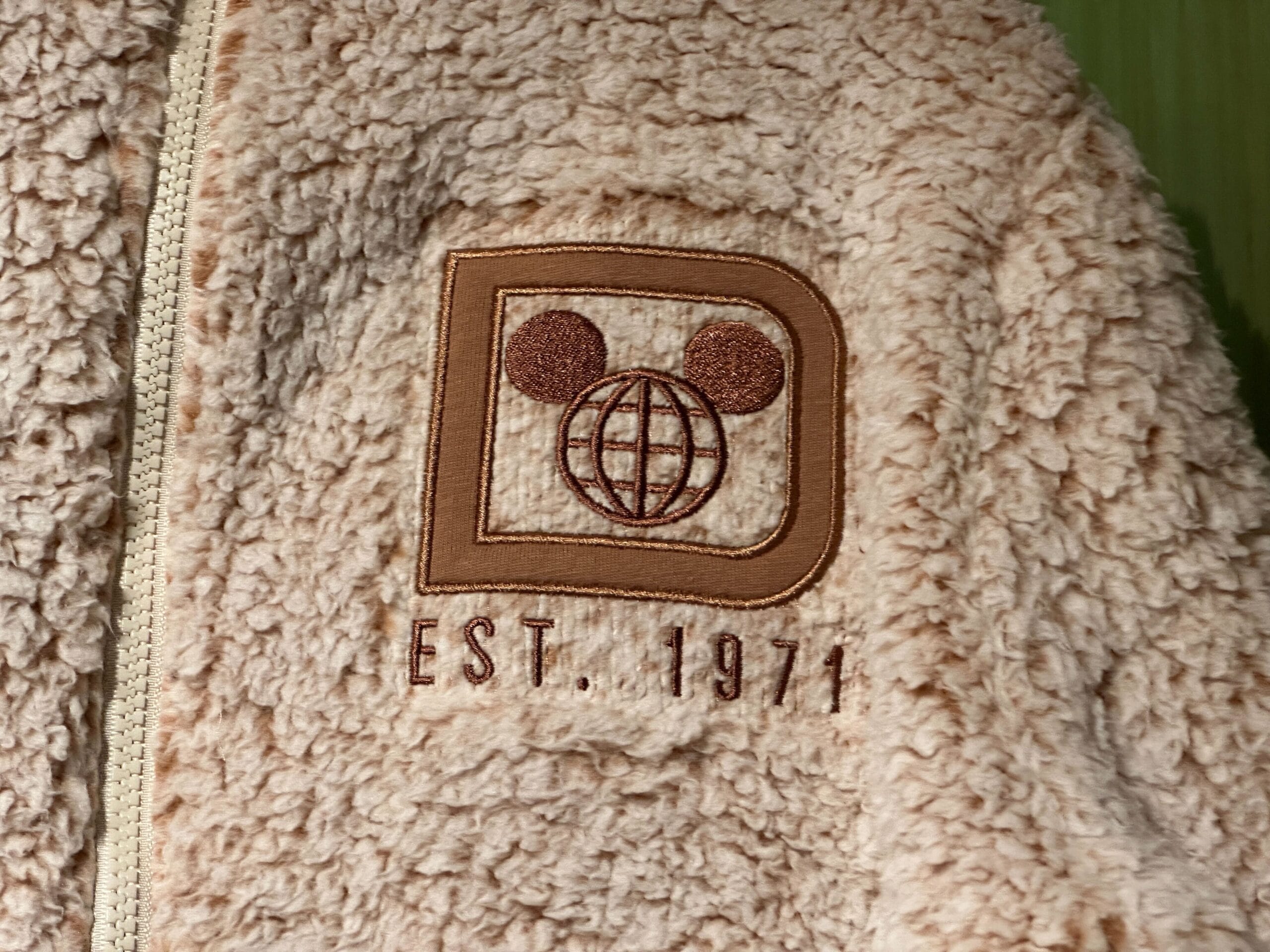 Beige fuzzy Authentic Original Jacket with zipper, Walt Disney World globe, Mickey ears, and "EST. 1971" patch, referencing Florida park.