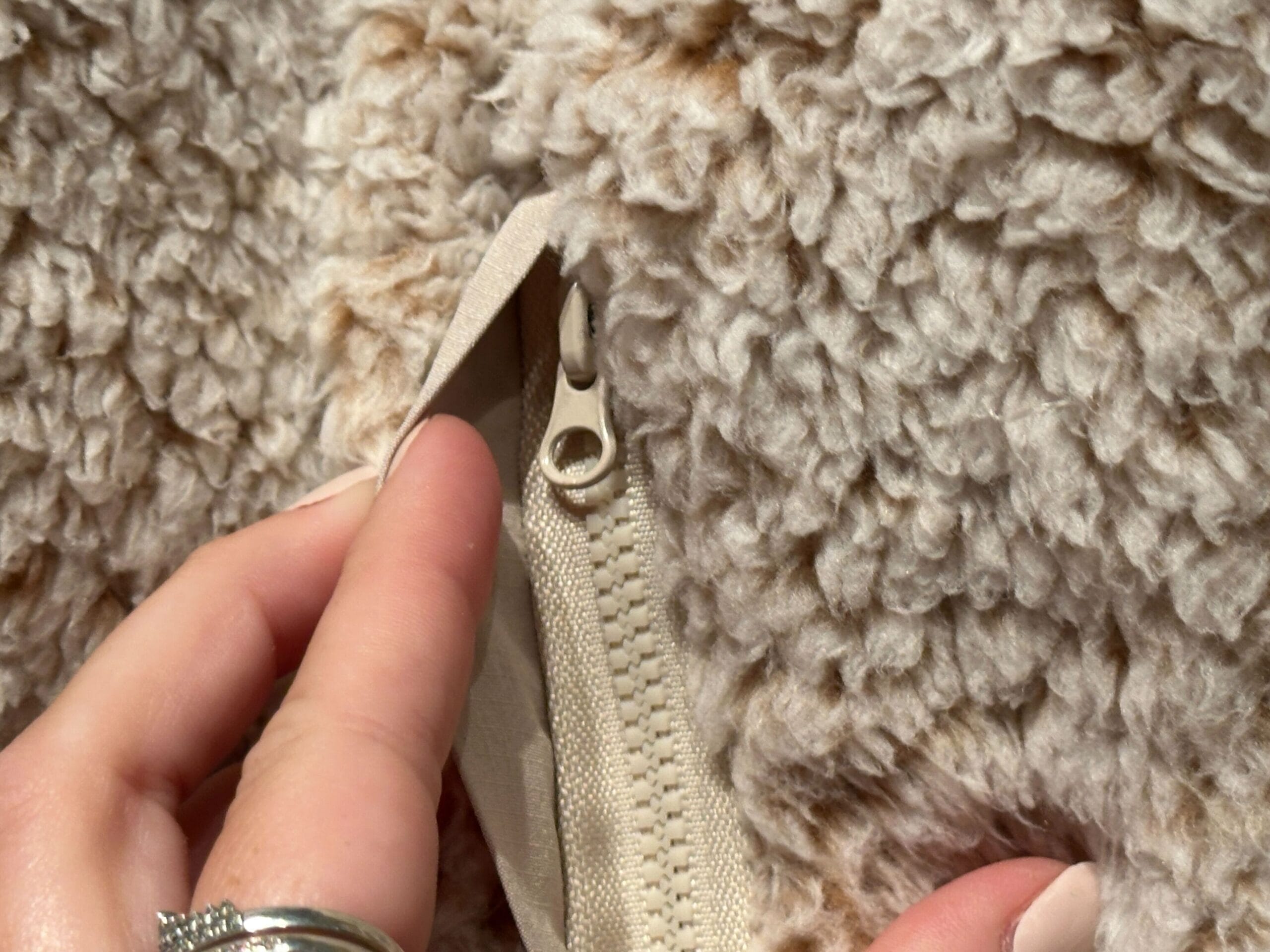 A hand unzips a beige zipper on a plush, cream jacket—cozy like a Disney spirit jersey, but no castle like Disneyland’s visible.