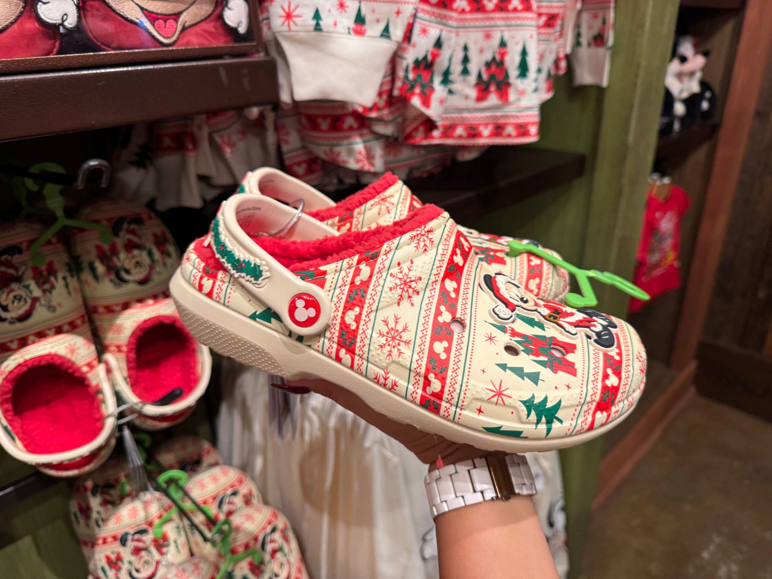 A hand holds up a festive Walt Disney World holiday clog shoe with Mickey Mouse and Christmas merchandise patterns.