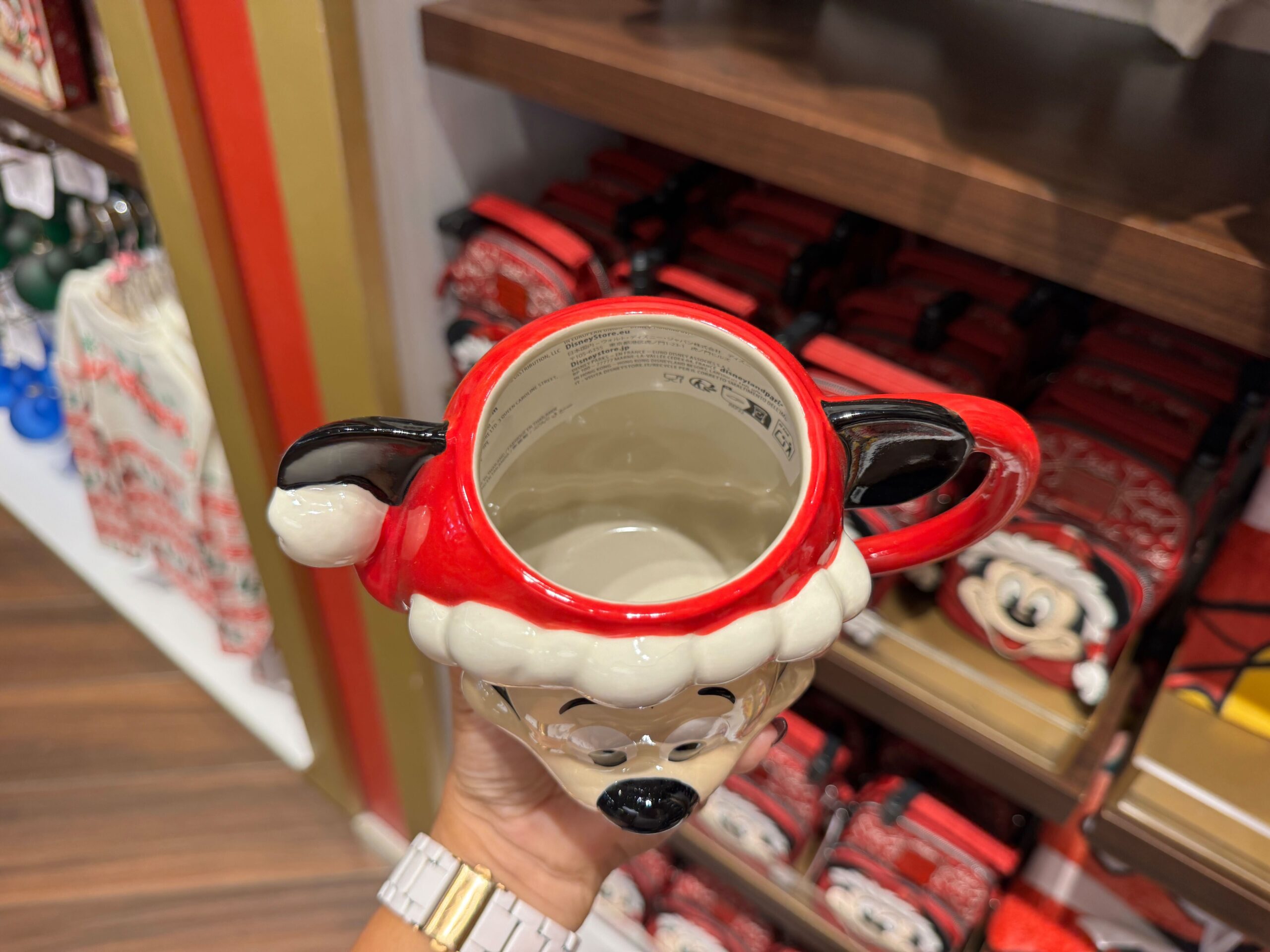 A hand holds a Santa Mickey Mouse mug, part of the 2025 Disney merchandise lining store shelves in the background.