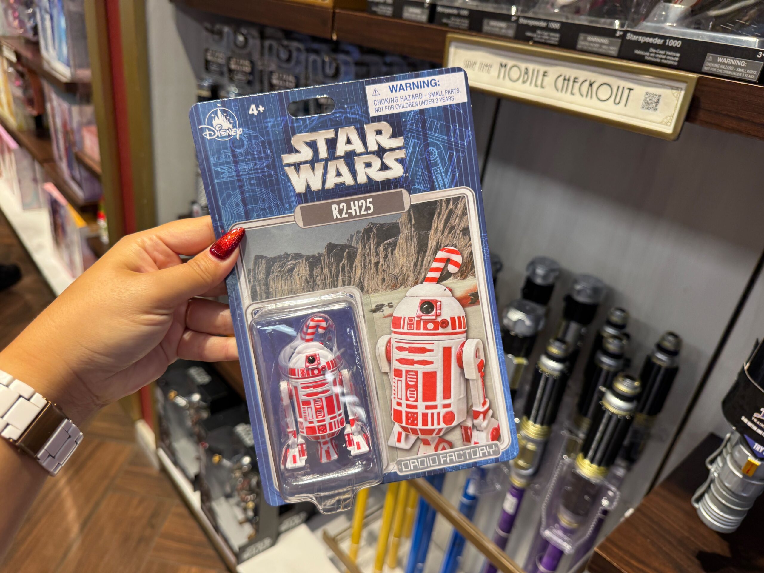 A hand holds a packaged red-and-white R2-H25 droid toy, 2025 Christmas merchandise at Walt Disney World, near display lightsabers.