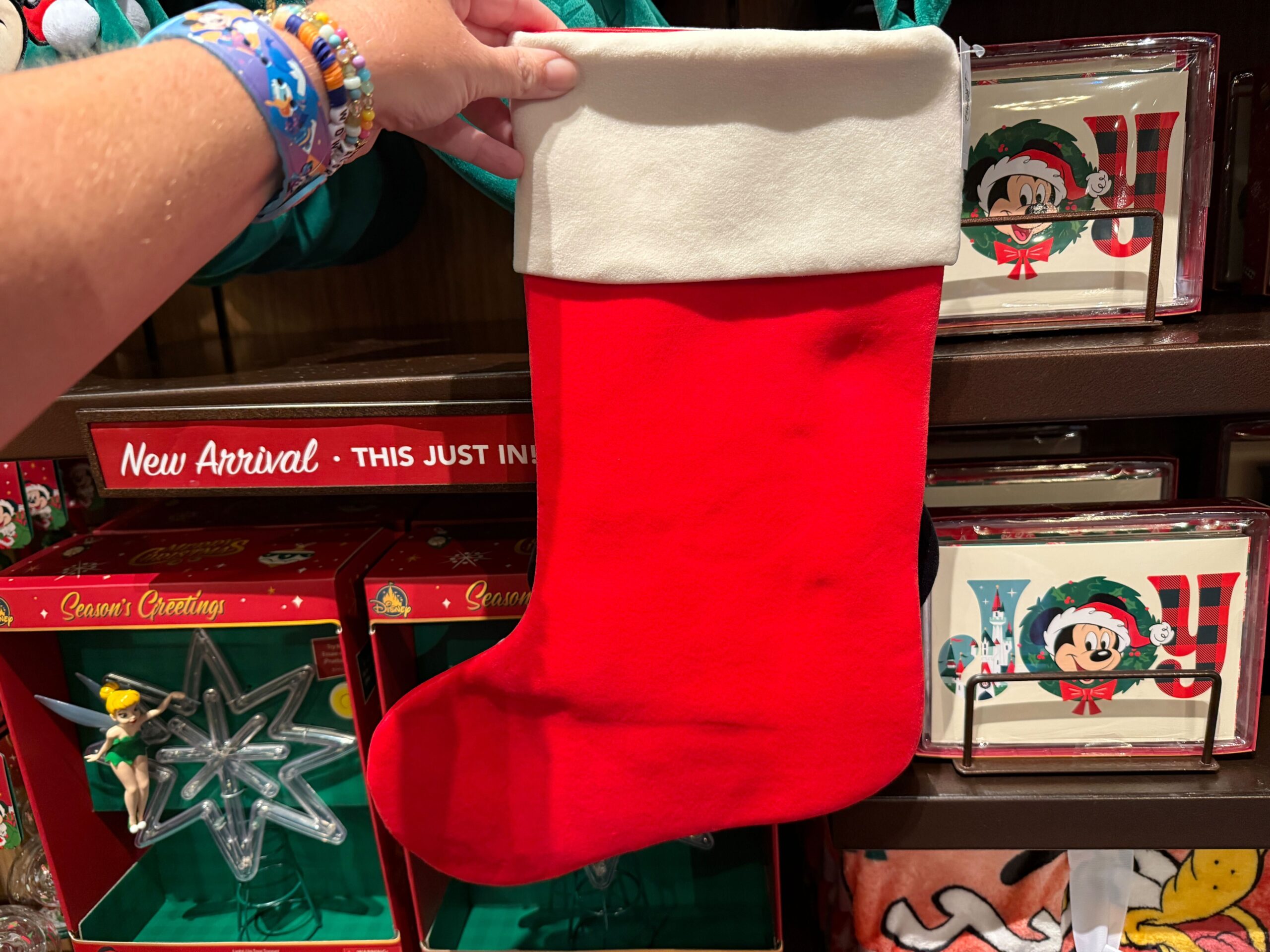A hand holds a plain red and white Christmas stocking in a store, with 2025 Walt Disney World holiday decor on shelves behind.