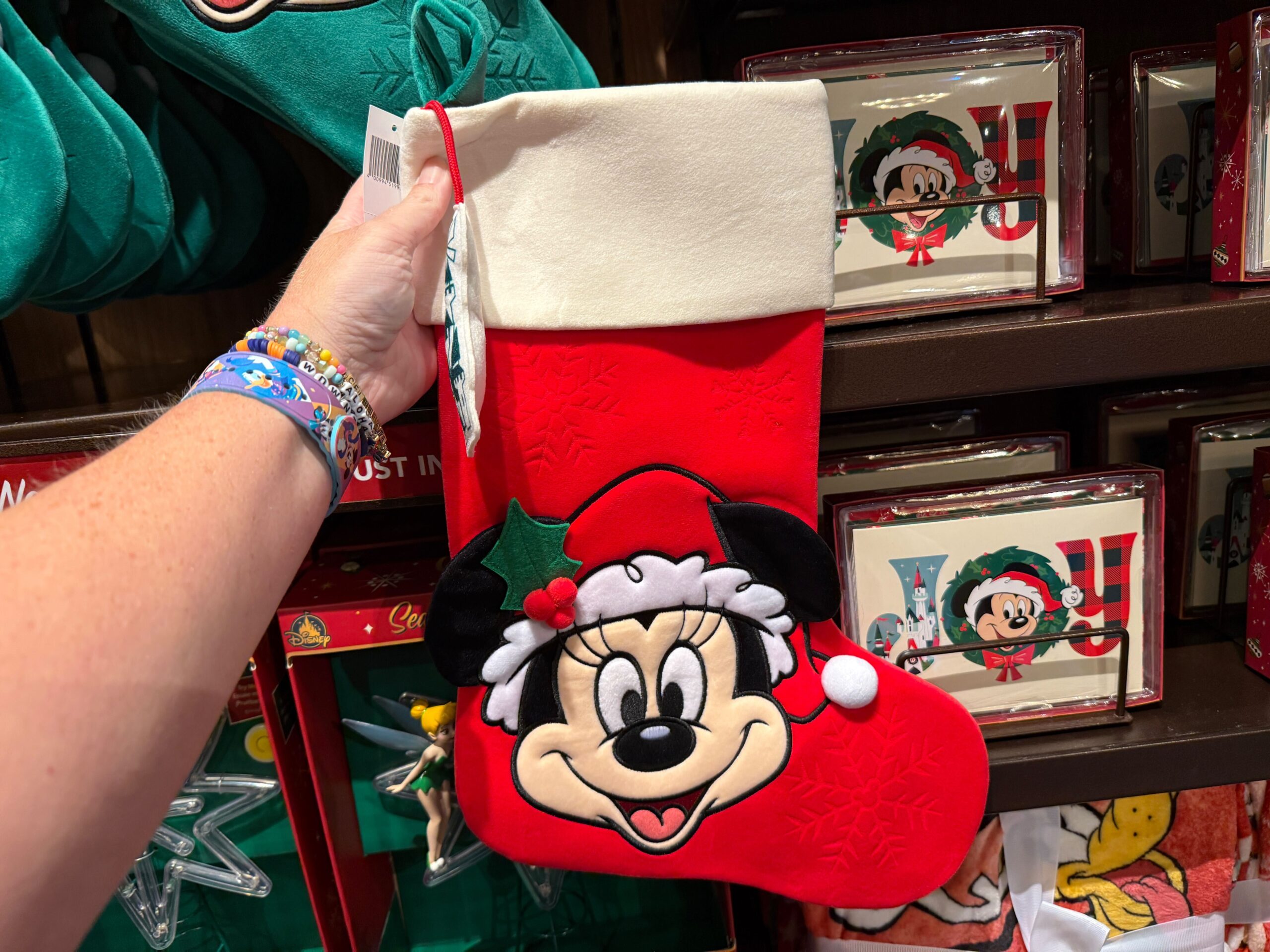 A hand holds a 2025 Minnie Mouse Christmas stocking in a Santa hat with holly, at a Walt Disney World holiday display.