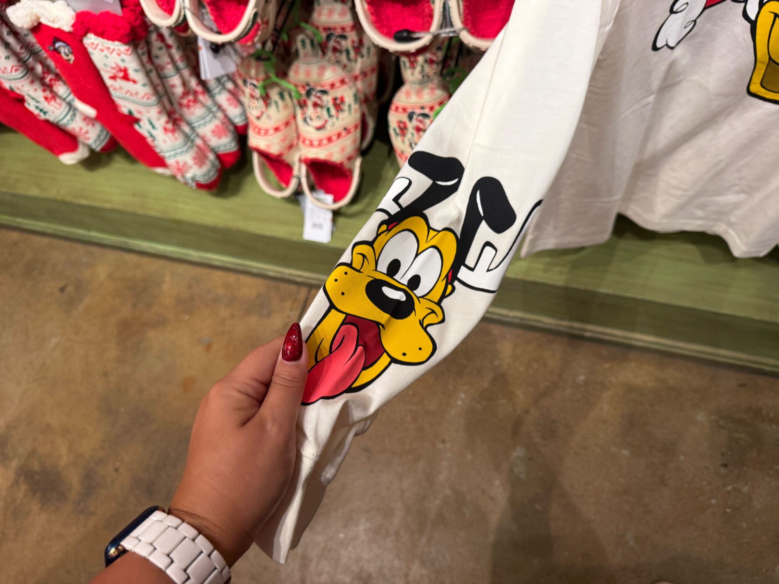 A hand holds a white shirt sleeve with a cartoon dog—Disney World Christmas merchandise for 2025—in a store setting.