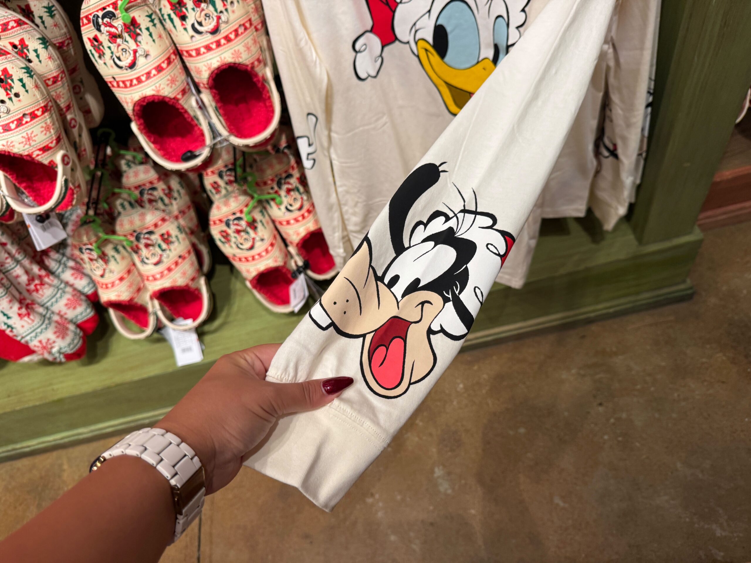 A hand holds a Goofy sleeve shirt; Christmas merchandise and festive shirts from Walt Disney World appear in the background.