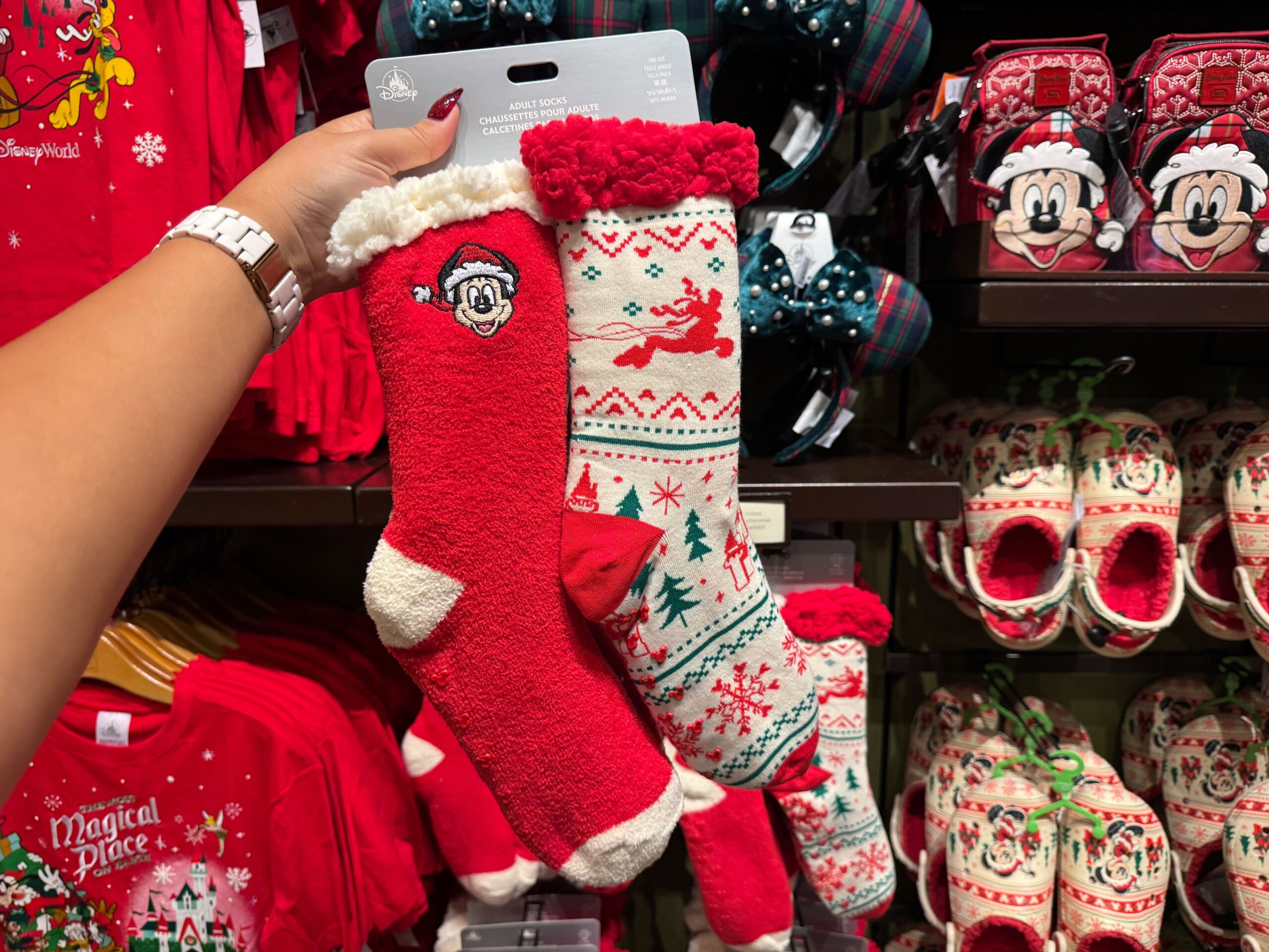 A hand holds two festive holiday socks amid Walt Disney World Christmas merchandise and clothing on display.