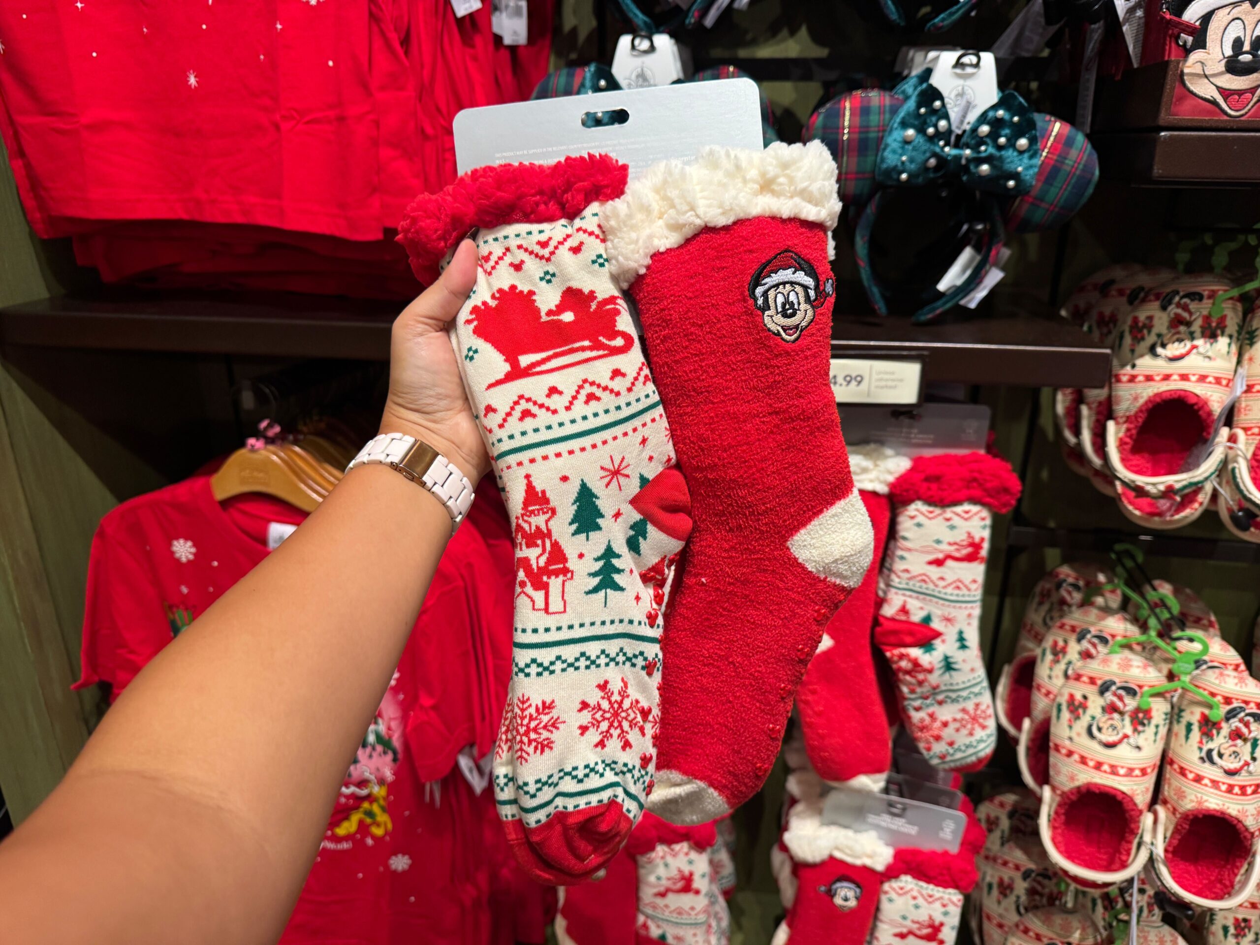 A hand holds two festive Walt Disney World Christmas socks in a store, surrounded by cheerful 2025 holiday clothing.