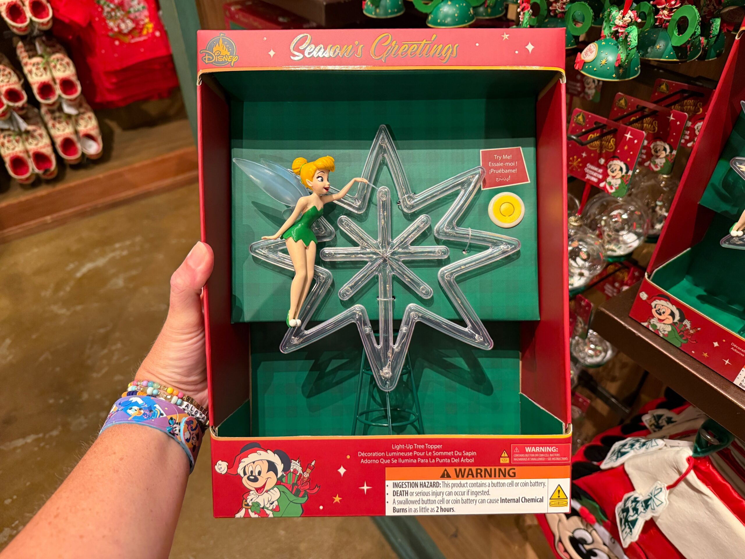 A hand holds a 2025 Tinker Bell light-up tree topper in holiday packaging with "Season's Greetings" text.