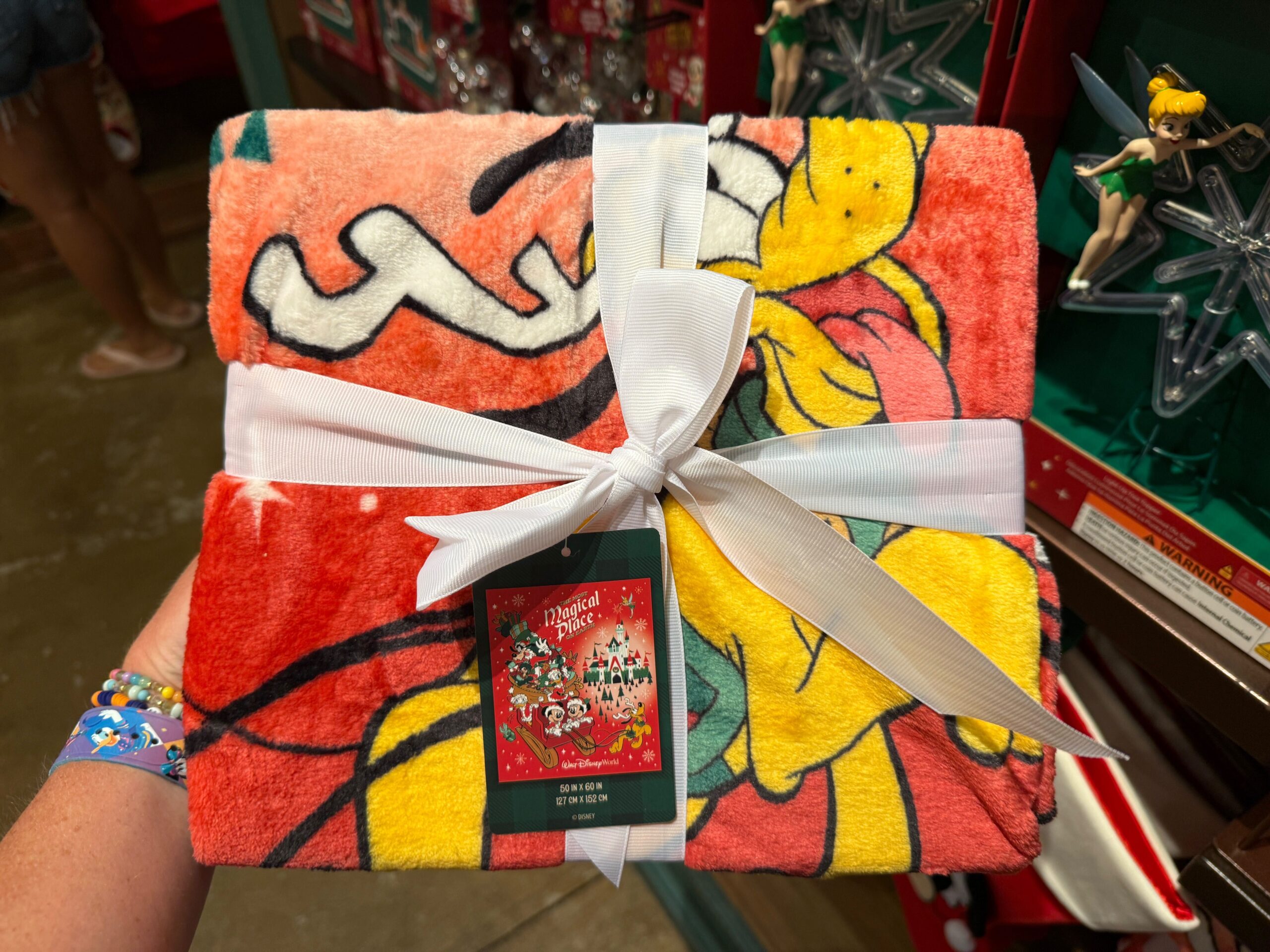 A hand holds a folded Winnie the Pooh holiday blanket—2025 Christmas merchandise—tied with a white ribbon and red tag.