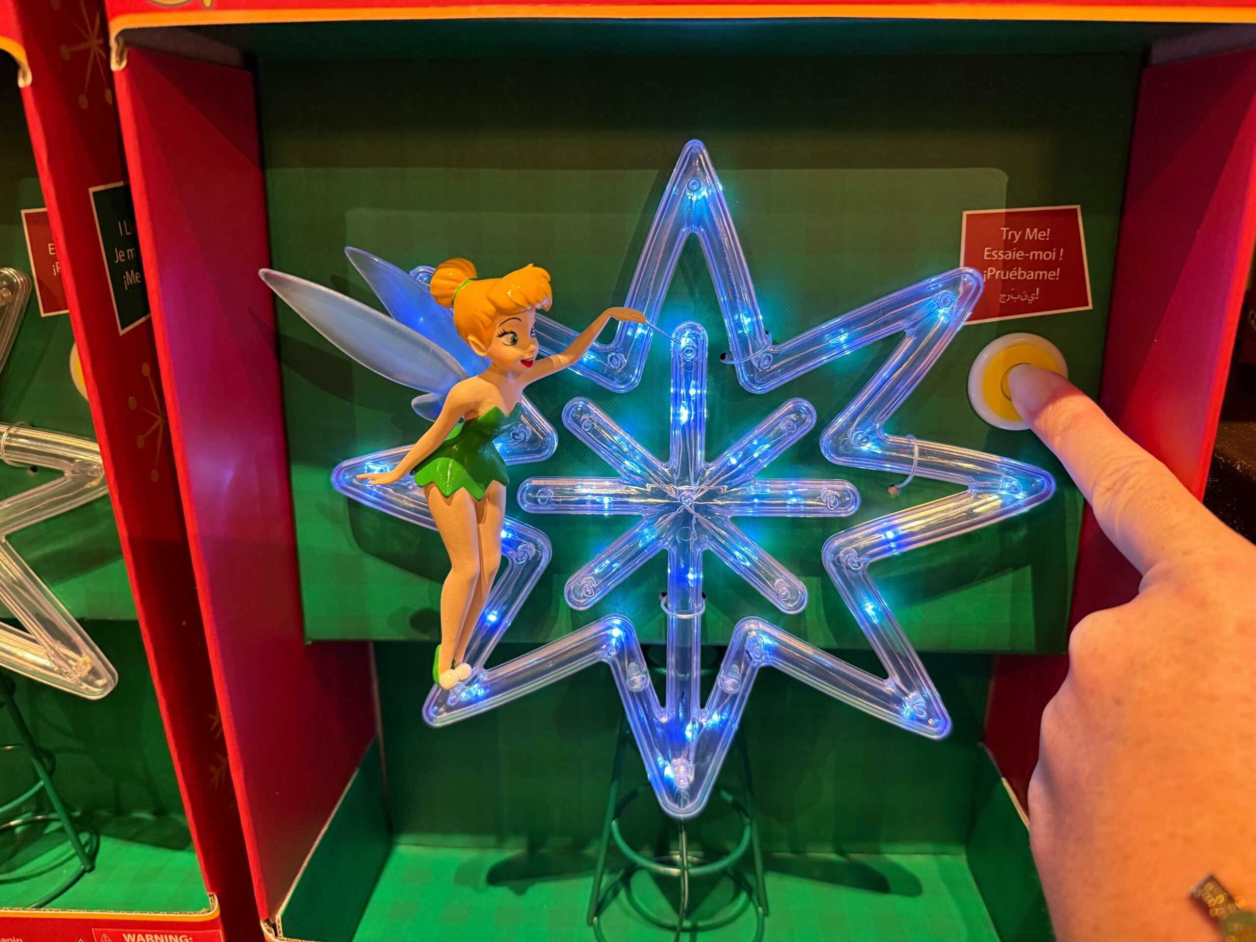 A Disney World Christmas merchandise Tinker Bell ornament glows blue as a hand presses the "Try Me!" button.