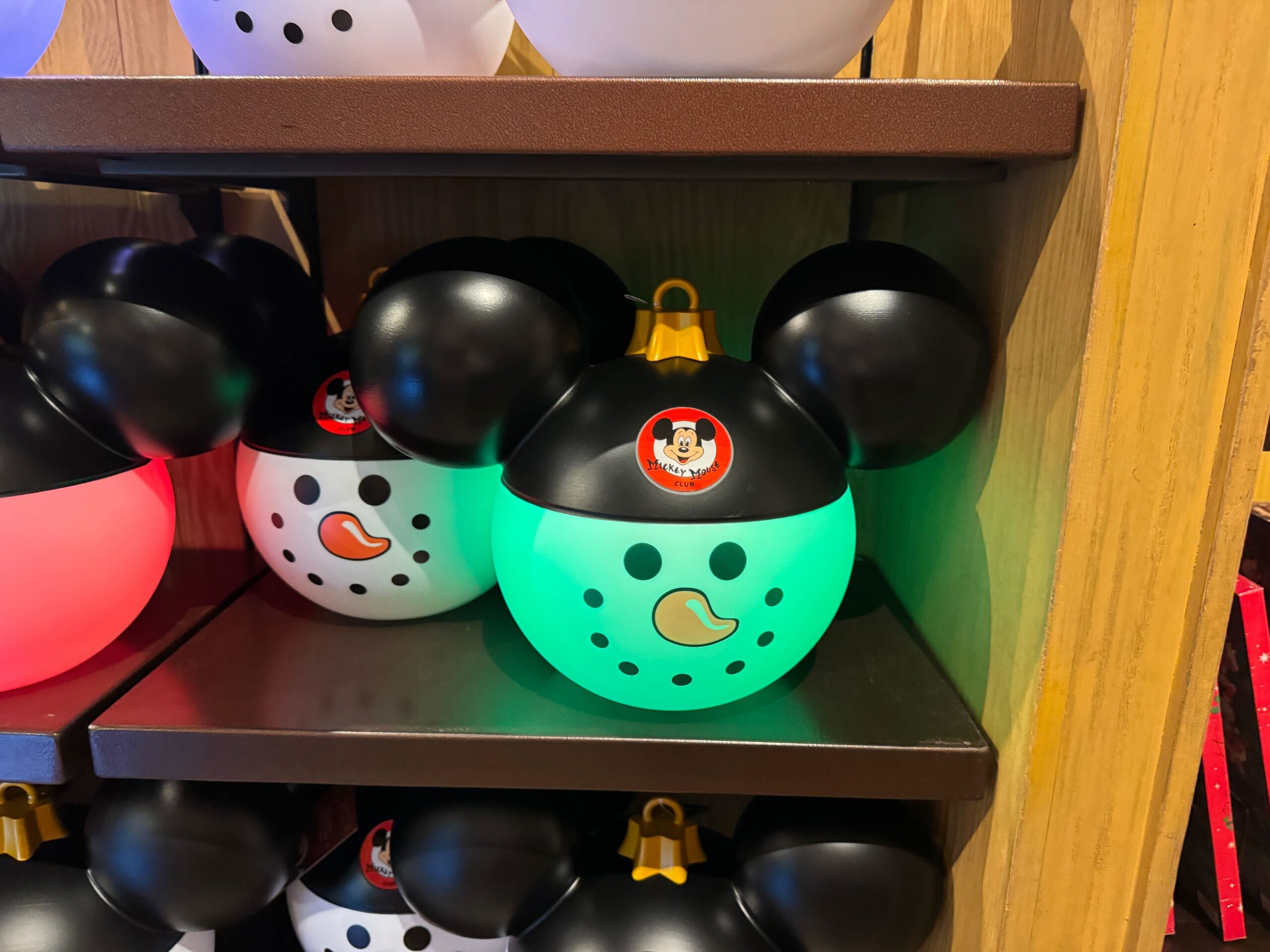 Green glowing Mickey Mouse snowman ornament, part of 2025 Walt Disney World Christmas merchandise, on a wooden shelf.