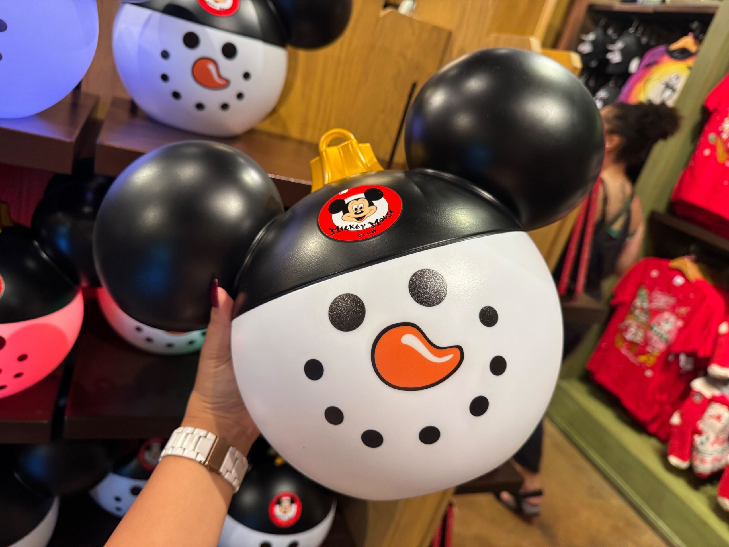 A hand holds a snowman-shaped Mickey Mouse popcorn bucket among 2025 Christmas merchandise at Walt Disney World.