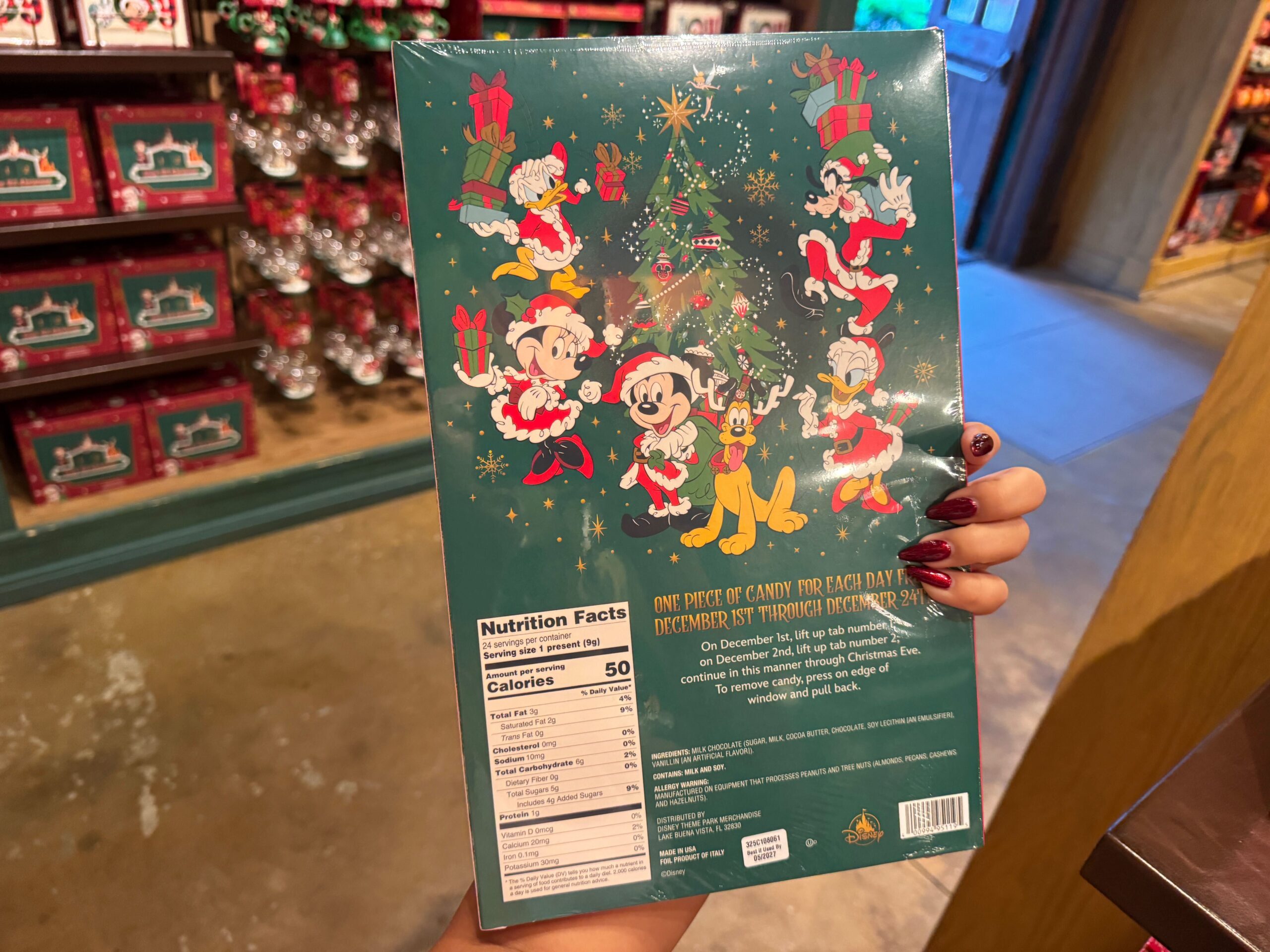 A hand holds Walt Disney World Christmas merchandise: a candy box with Mickey, Minnie, Goofy, Pluto, and Donald in festive outfits.