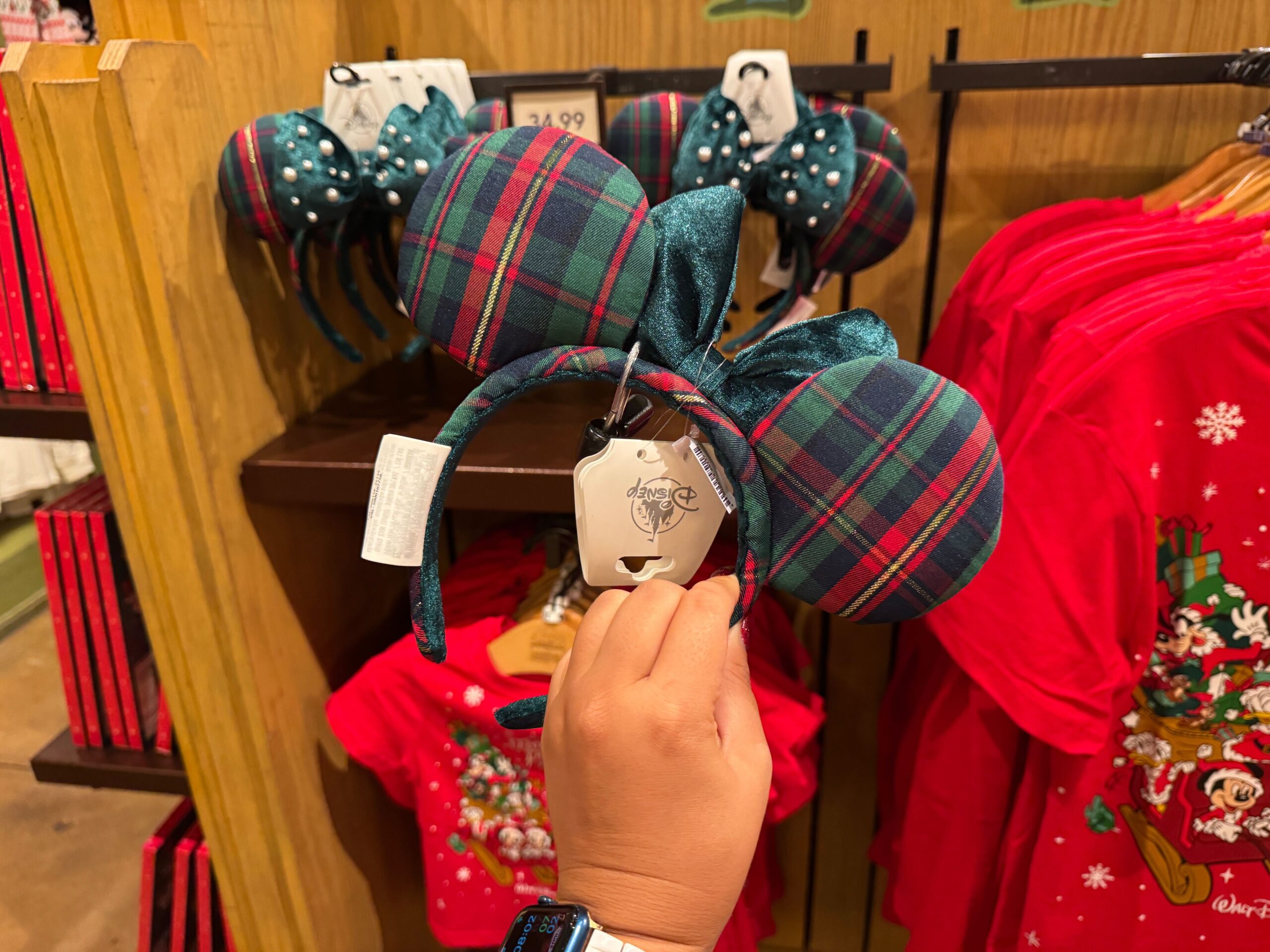 A hand holds up plaid Minnie Mouse ears among 2025 Walt Disney World Christmas merchandise and festive shirts on display.