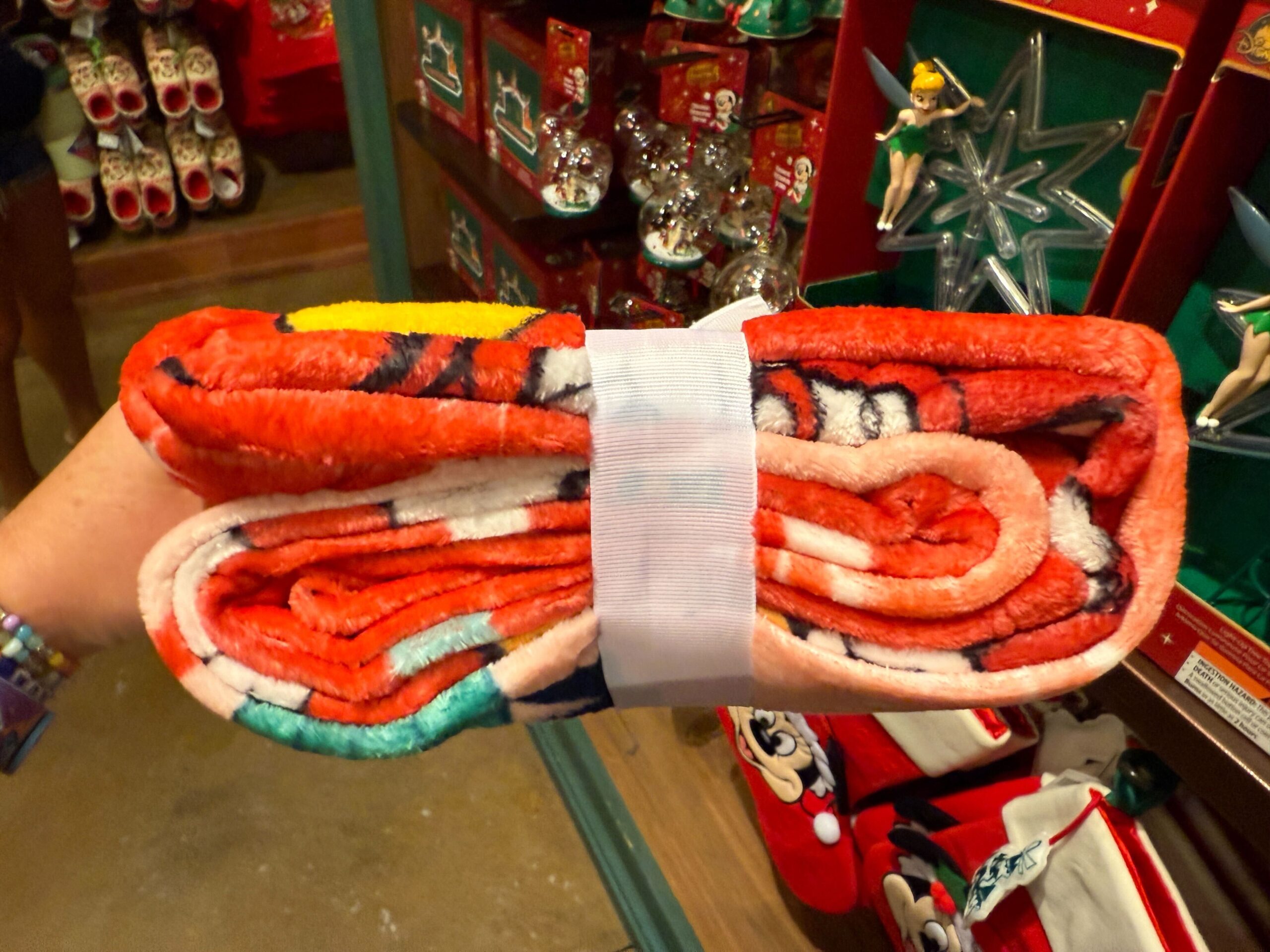 A hand holds a folded, colorful plush blanket among Walt Disney World Christmas merchandise in the background.