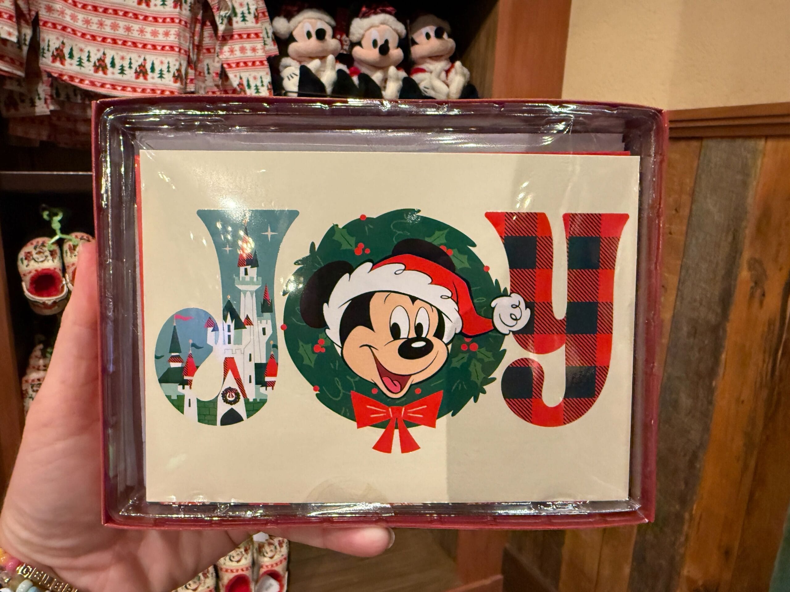 A hand holds a "JOY" card with a castle and Mickey Mouse, part of Walt Disney World Christmas merchandise in a festive display.