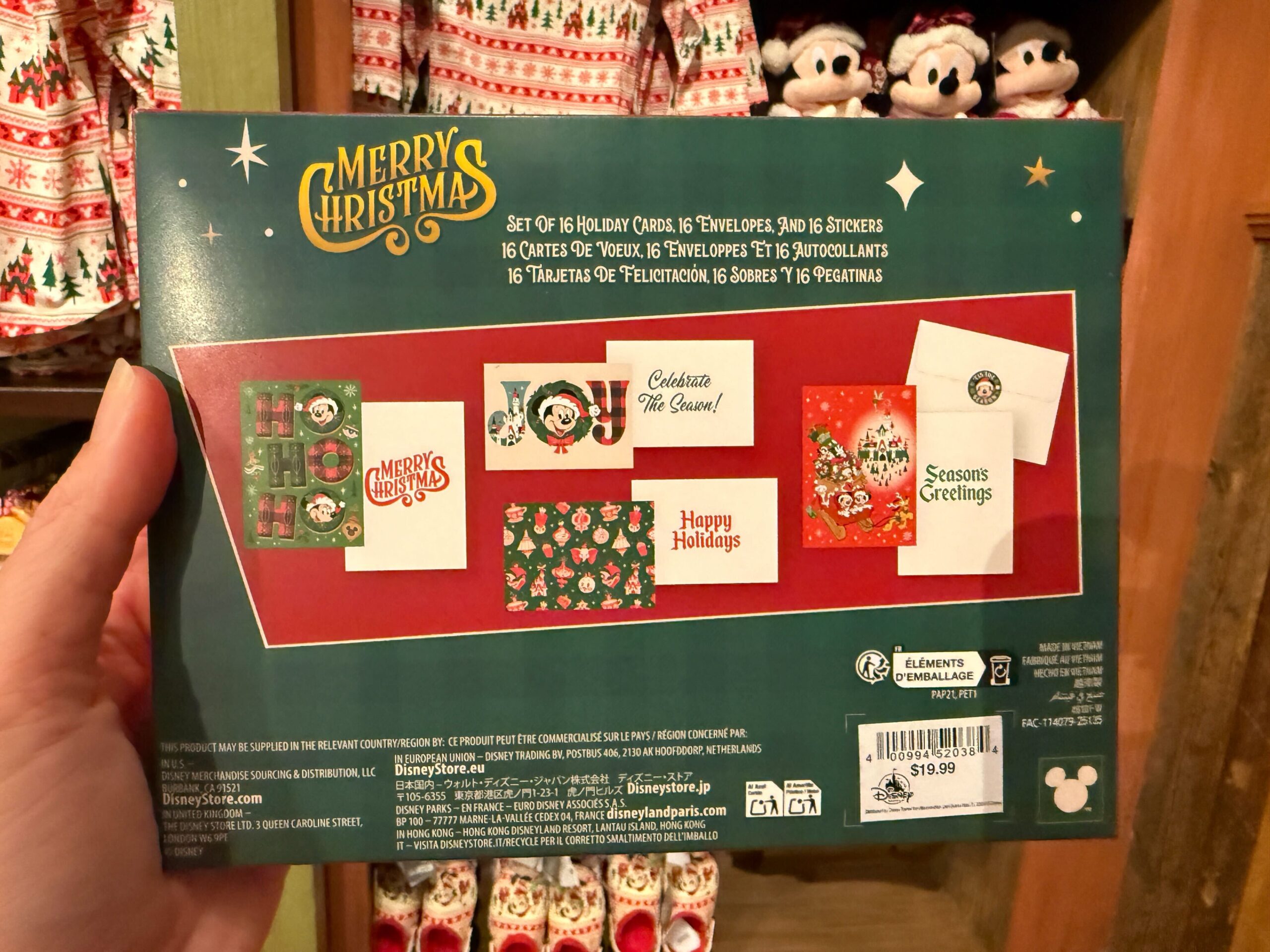 A hand holds a box of Walt Disney World Christmas merchandise—cards, envelopes, and stickers—in a festively decorated store.