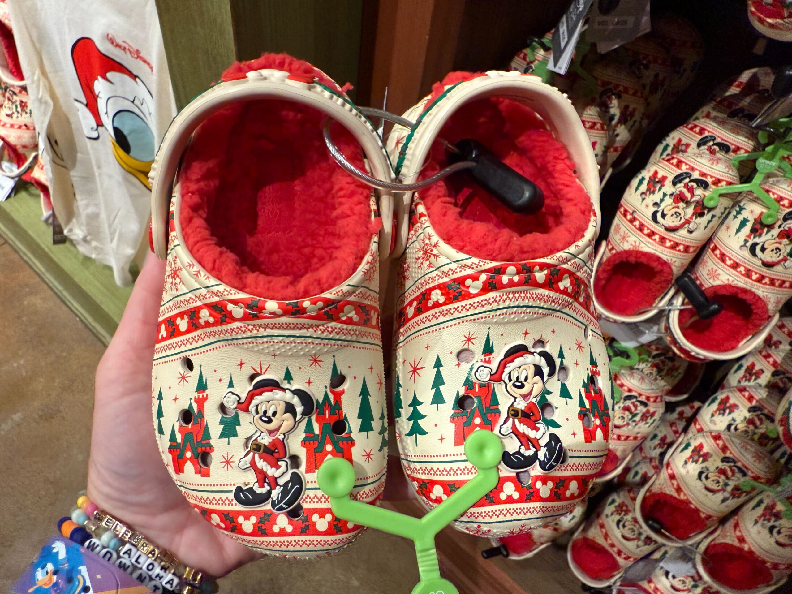 A hand holds festive Walt Disney World holiday clogs featuring Mickey Mouse in a Santa hat, Christmas trees, and red interiors.