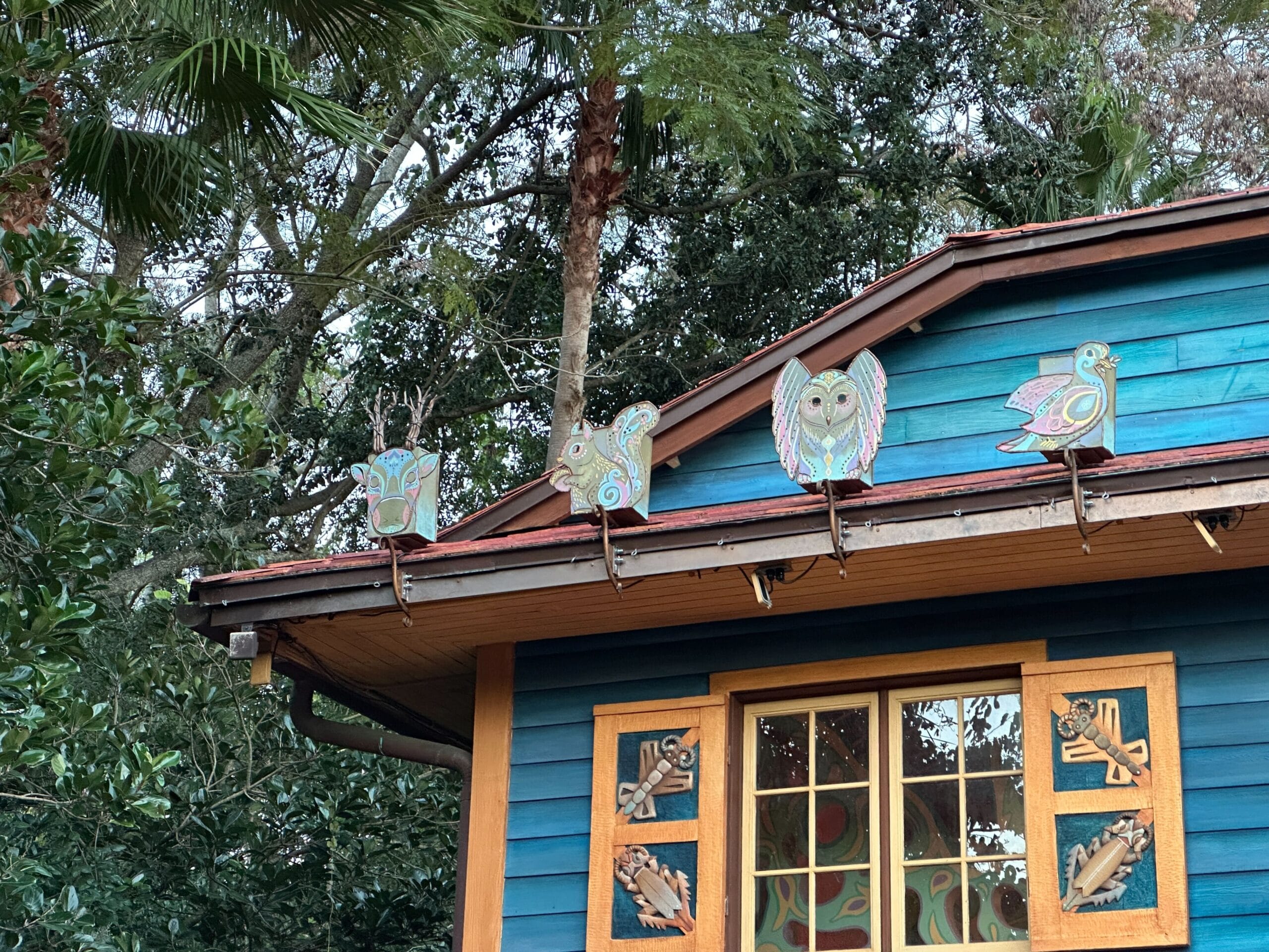Four vibrant bird sculptures bring holiday cheer to a blue house with ornate window carvings, echoing Disney’s festive park decor.