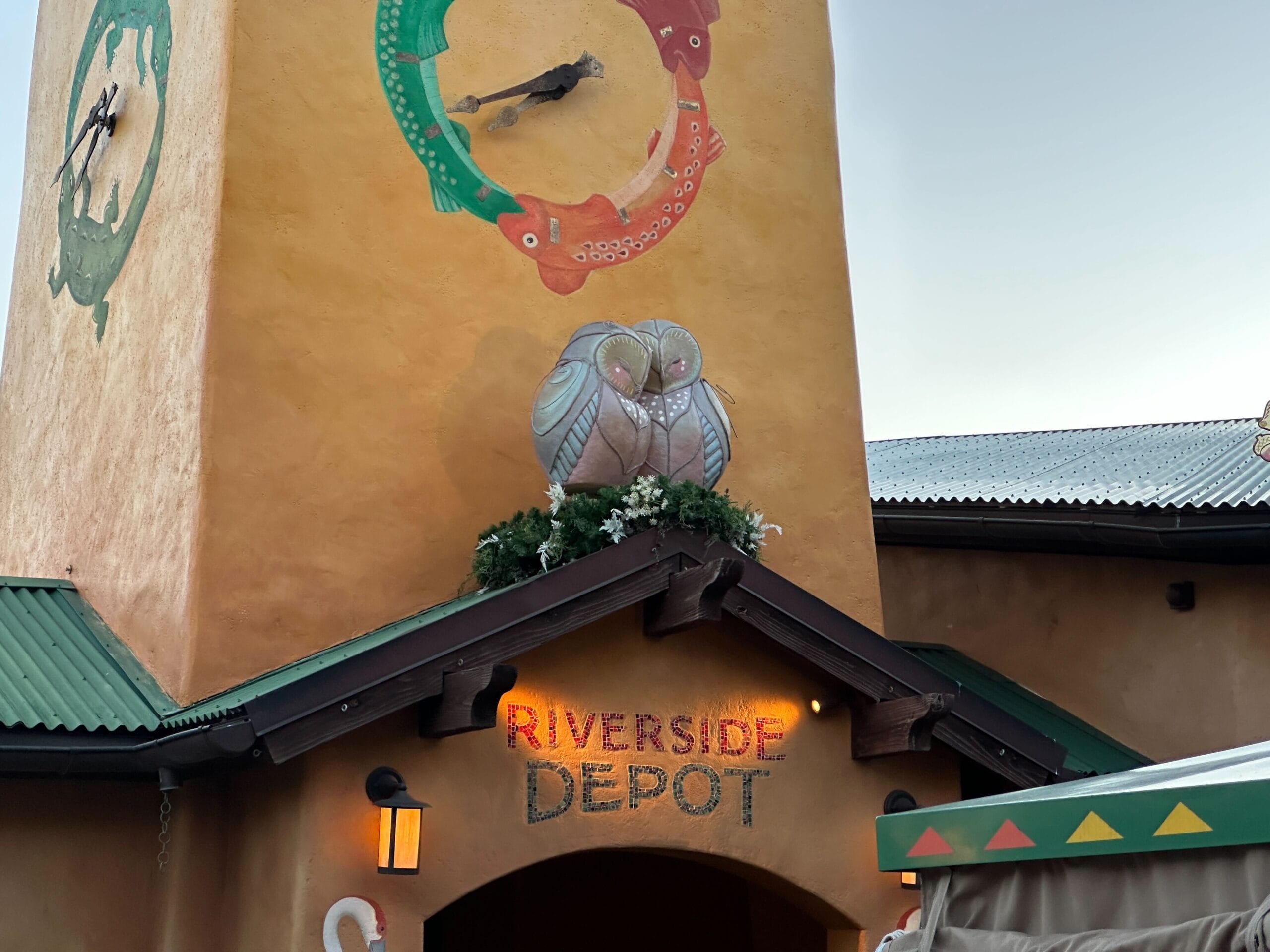 Riverside Depot at Animal Kingdom glows at dusk with festive Christmas decor, an owl statue, and a vibrant clock—no castle in sight.