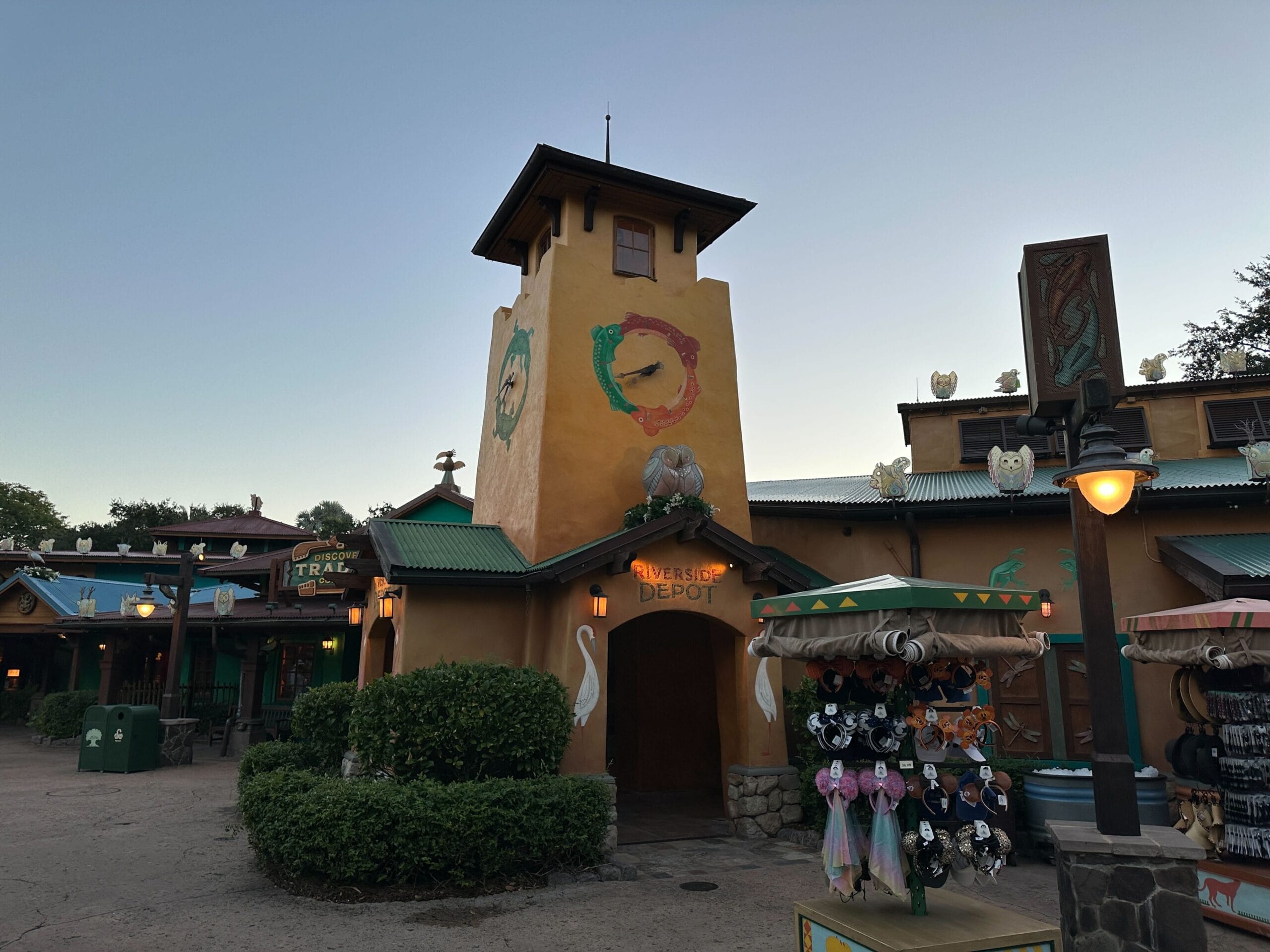 The Riverside Depot at Disney's Animal Kingdom, a tan building with animal murals and greenery, displays park merchandise outside.