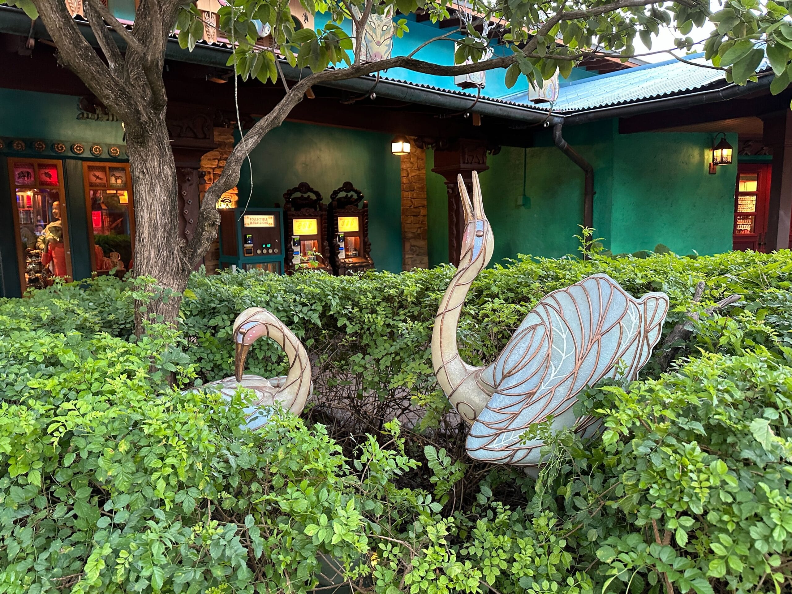 Two mosaic swan sculptures bring holiday cheer to bushes by a teal building with red-framed windows, evoking Disney park decor.