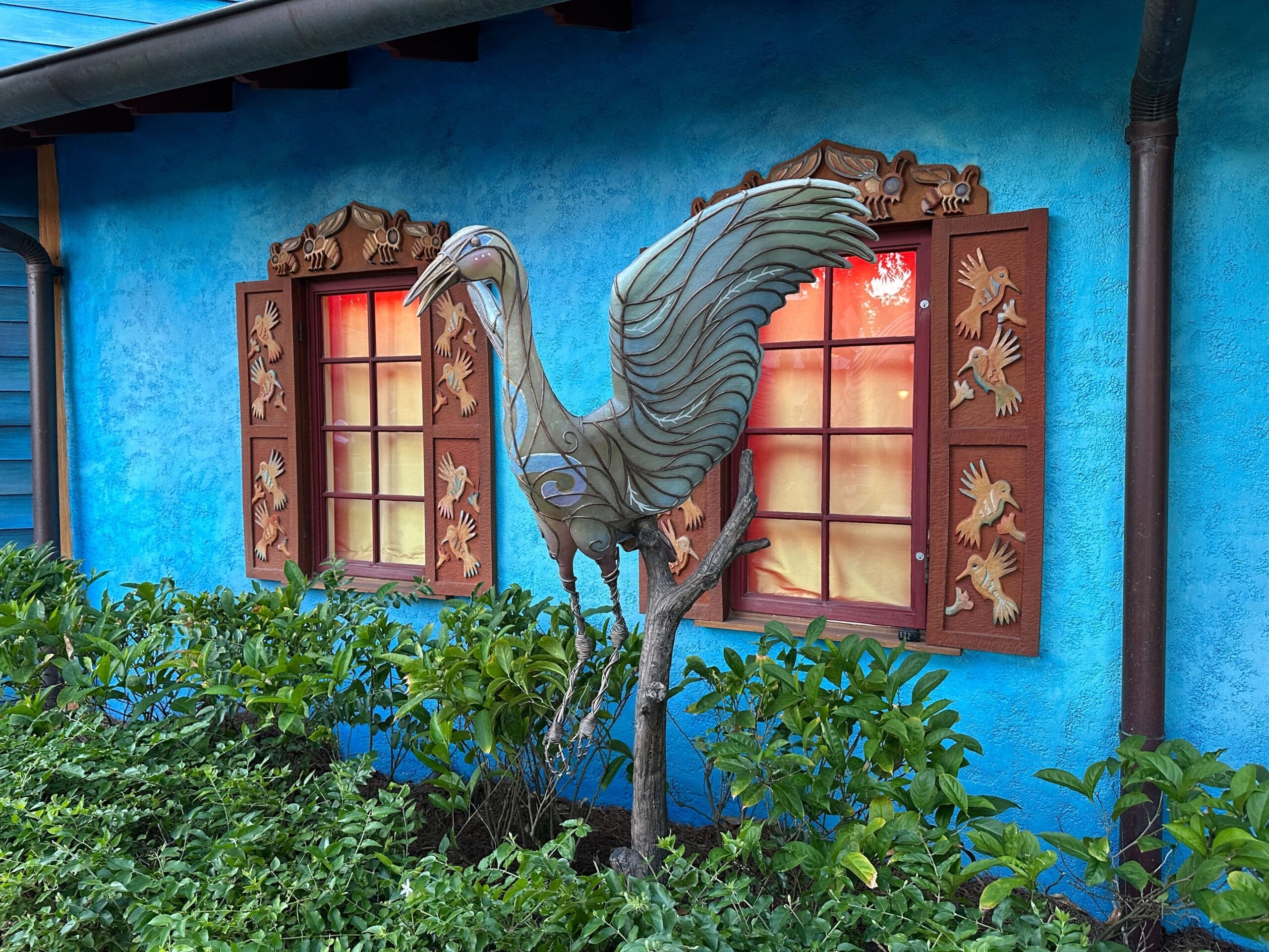 Metal bird sculpture with wings raised, in front of blue wall and windows with festive, Disney-style holiday shutters.