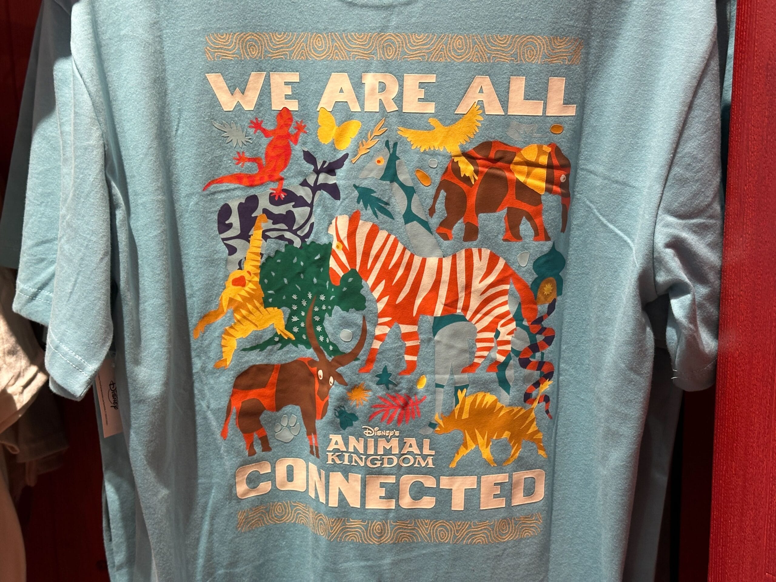 Light blue Animal Kingdom tee with vibrant animals, "We Are All Connected" and park logo; classic Disney Parks merchandise style.