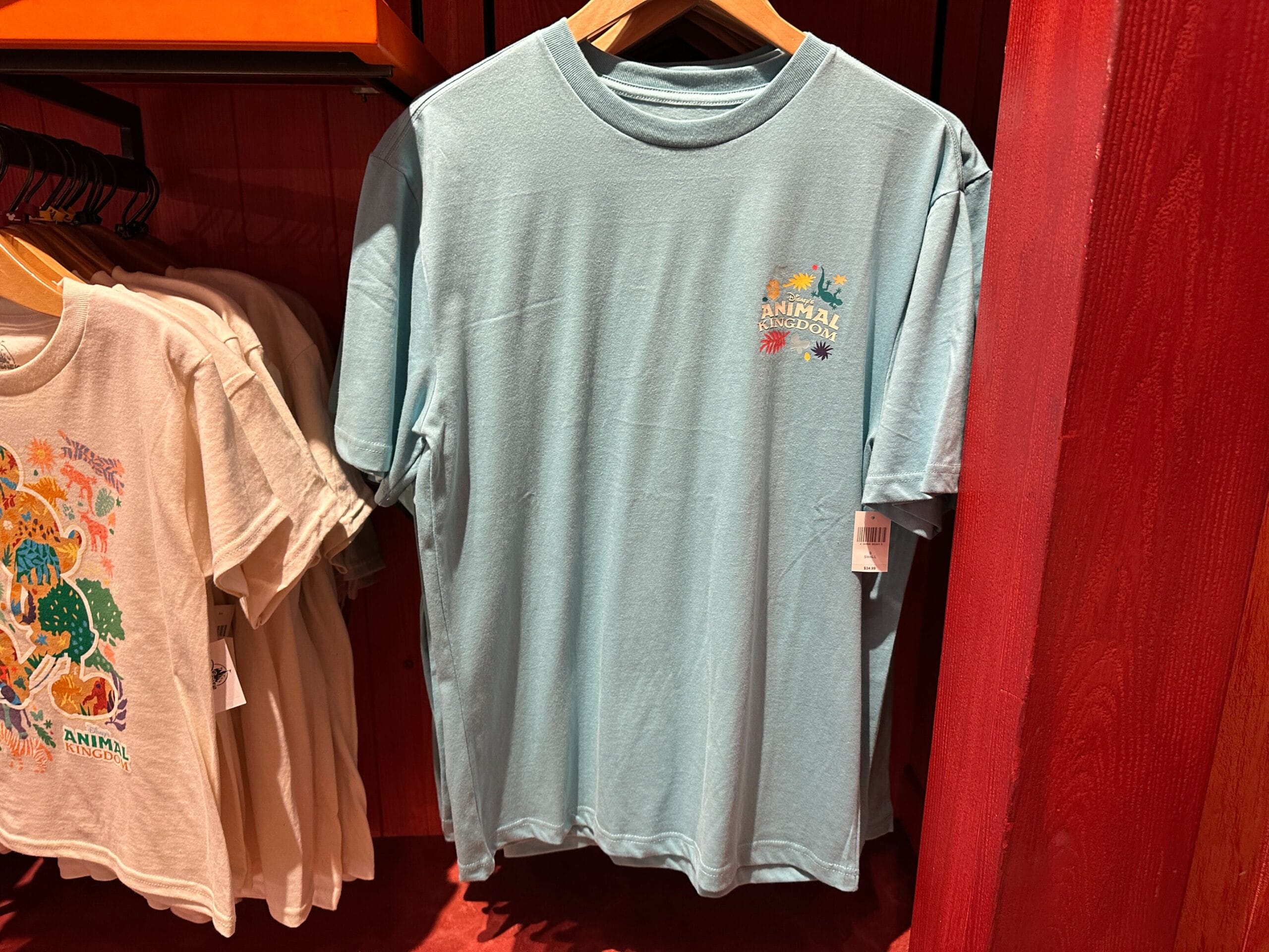 Light blue Disney tee with small Animal Kingdom logo, Circle of Life inspired, hanging on rack at a Disney park shop.